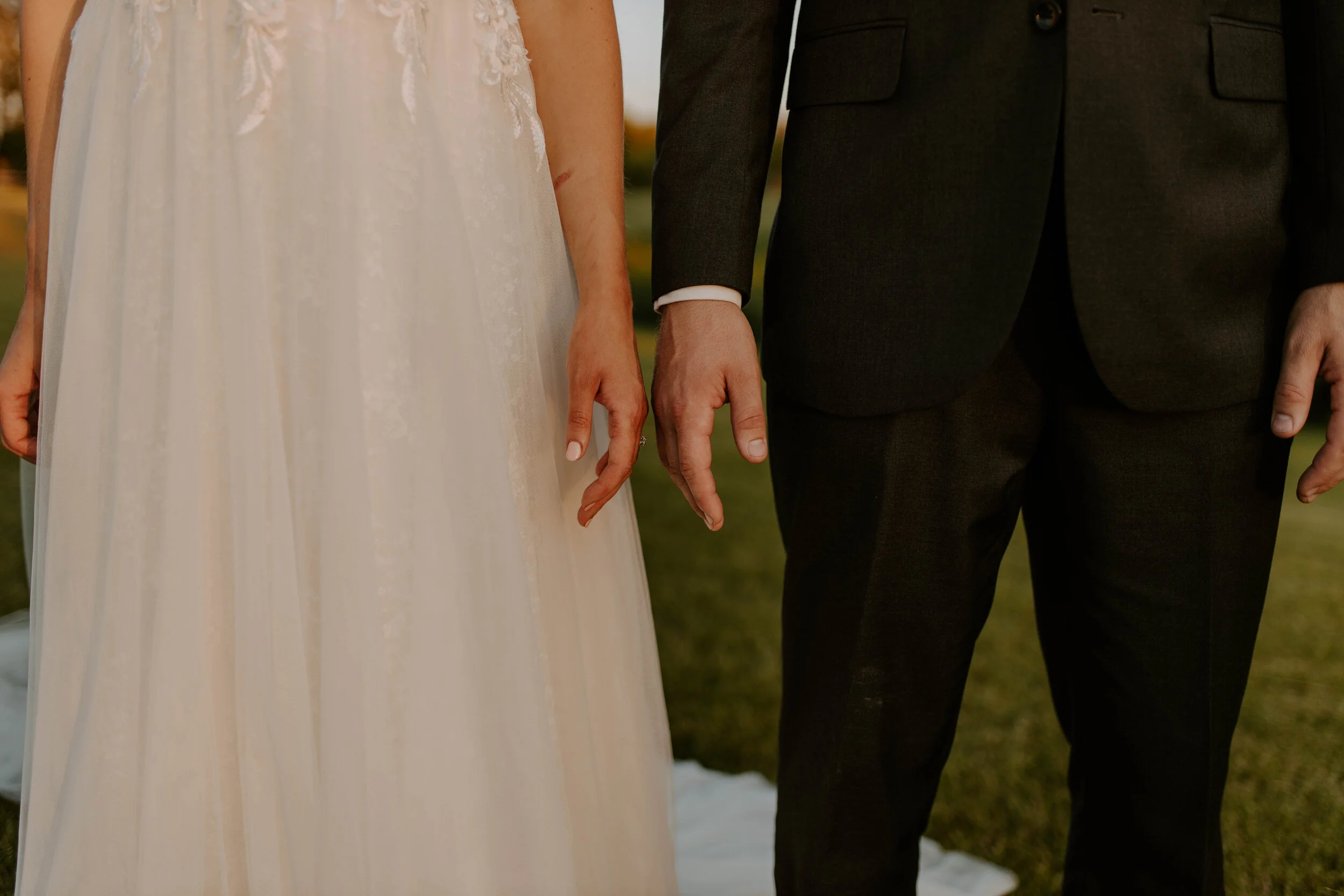 Close-up of a bride and groom holding hands during their wedding ceremony. The bride is wearing a white wedding dress with lace details, and the groom is in a dark suit.