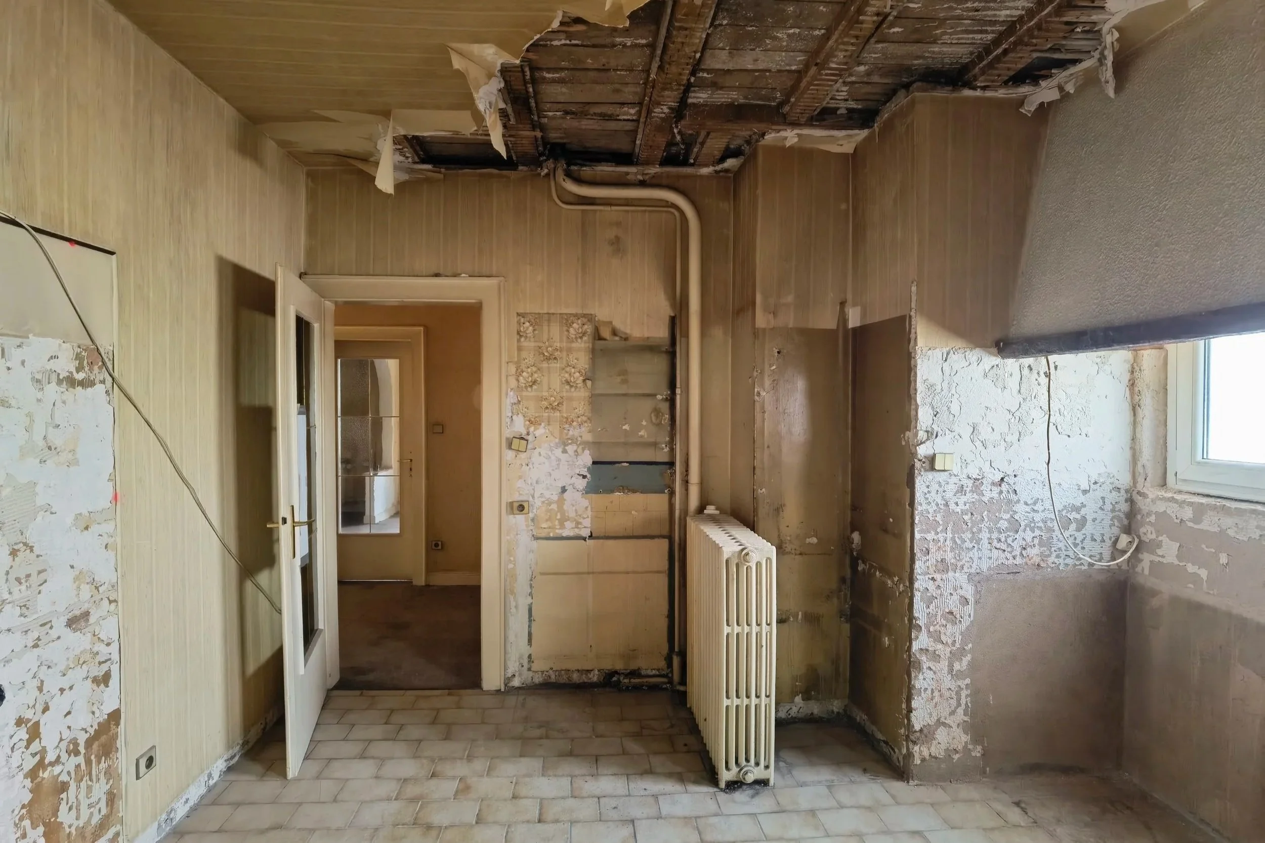 Salle de cuisine en rénovation, murs en mauvais état, plancher en carrelage, radiateur ancien, fenêtre, porte en verre, plafond en partie dégradé.