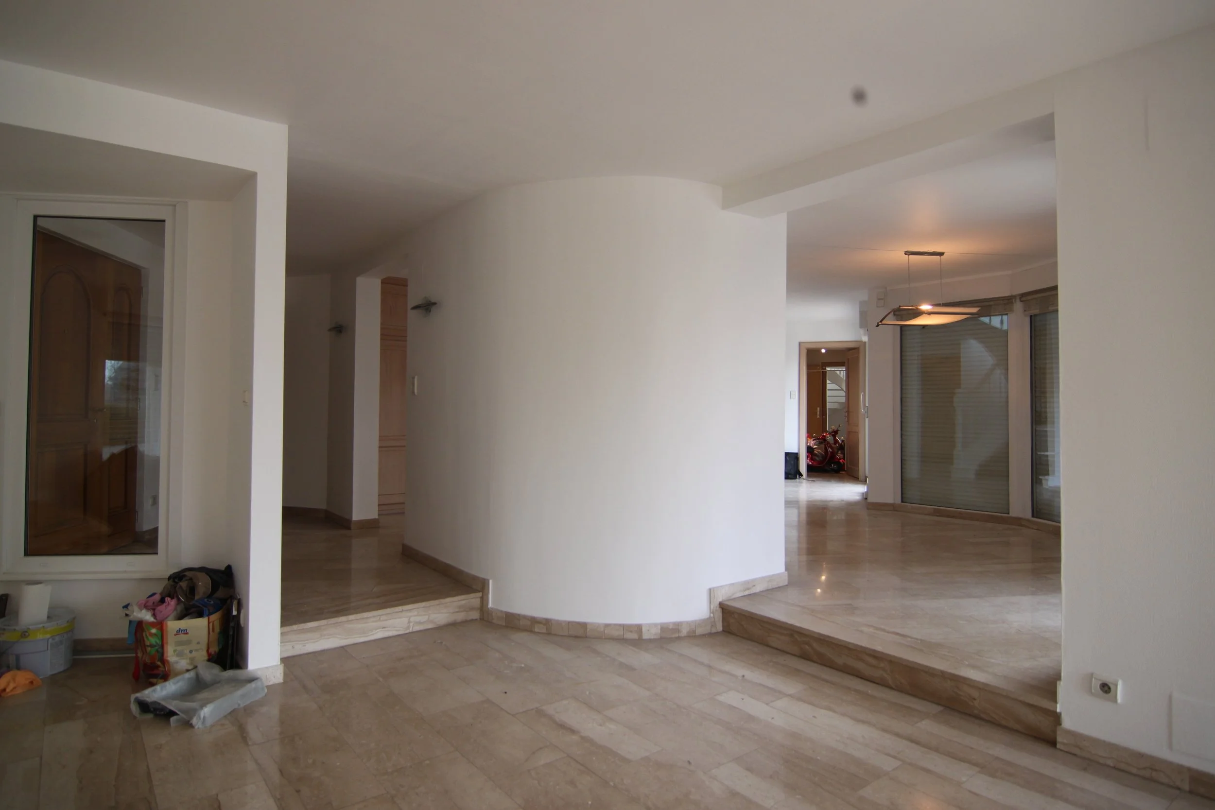 Intérieur d'une maison avec sol en marbre, murs blancs, escalier en basalte beige, avec un espace de vie spacieux et lumineux, quelques objets et un scooter visible à l'arrière.