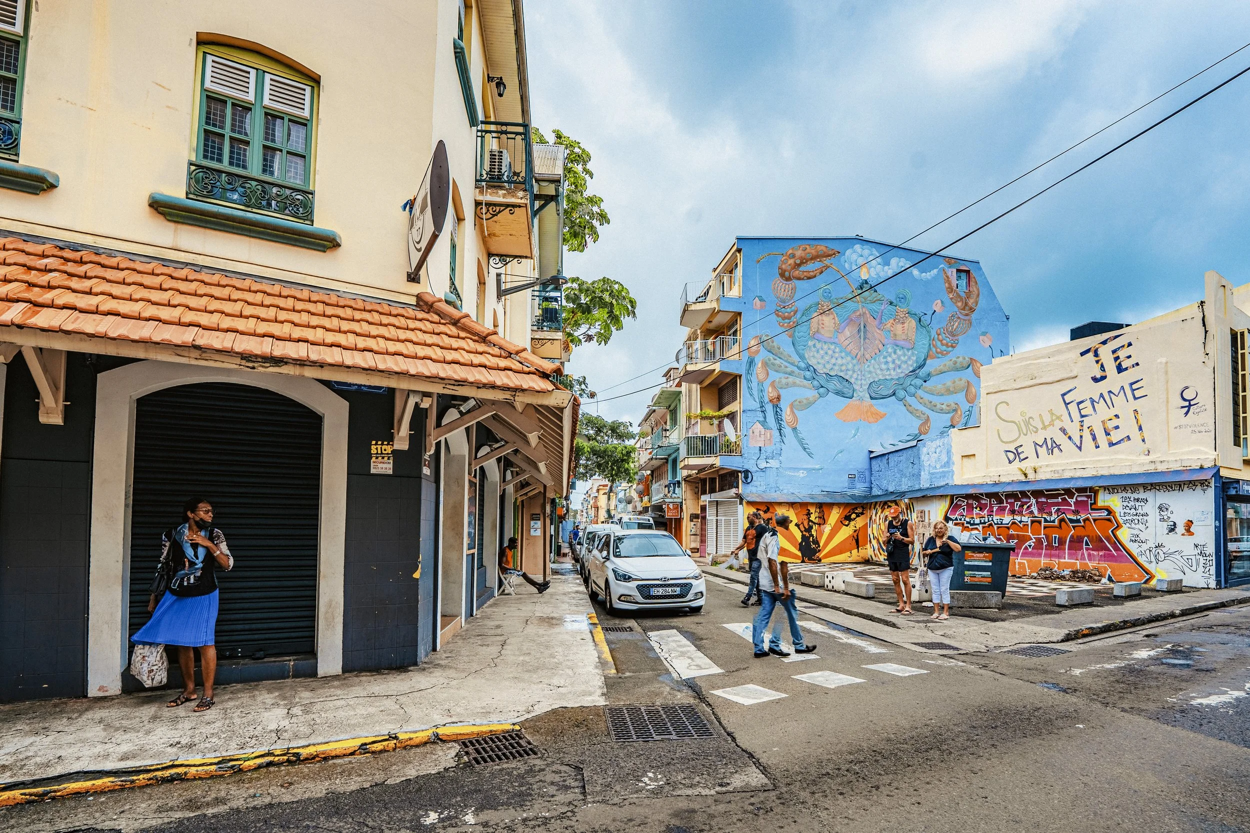 Rue animée avec des voitures garées, des piétons, et plusieurs bâtiments colorés en arrière-plan.