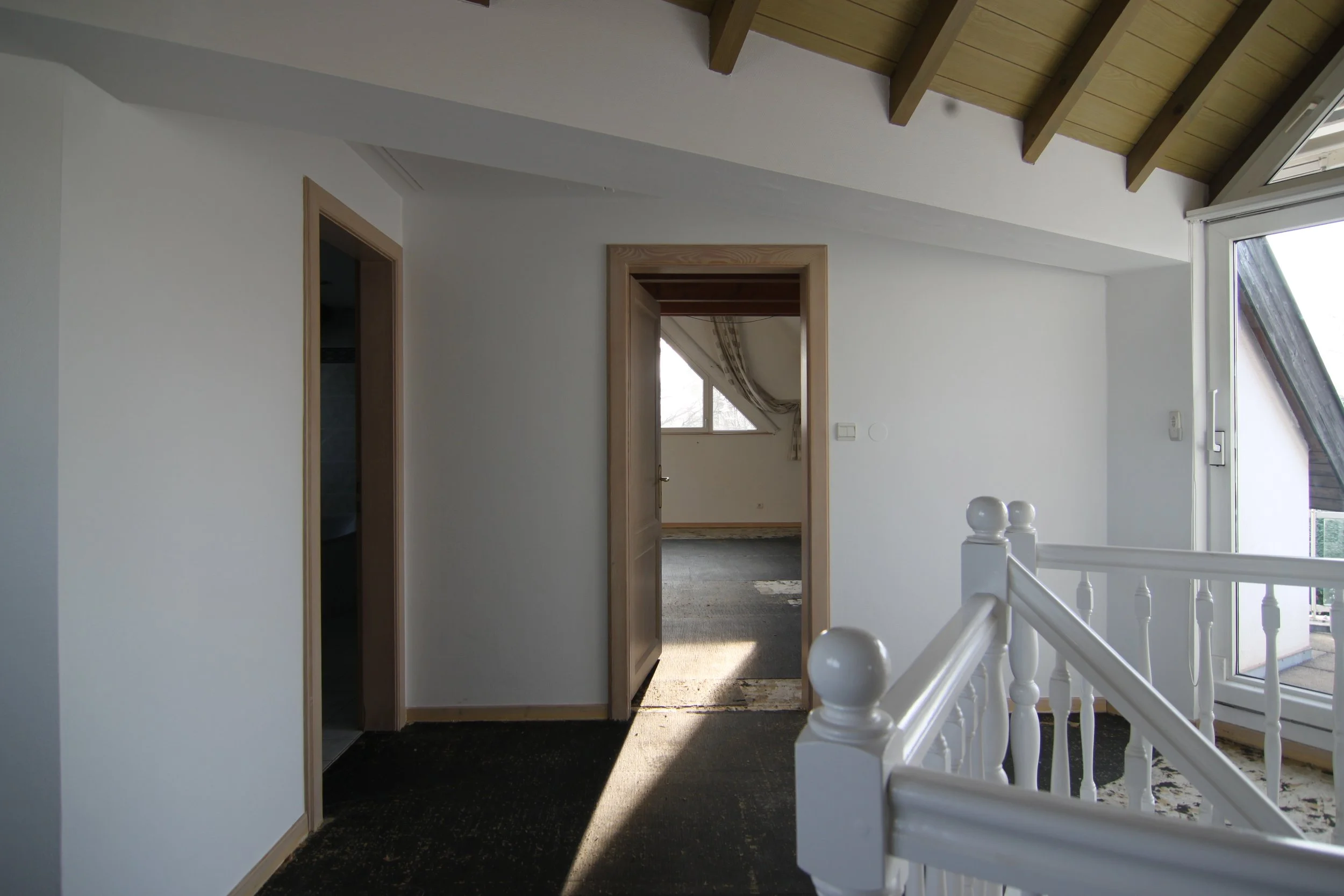 Intérieur d'une maison avec une pièce lumineuse, un escalier en bois peint blanc et deux portes menant à d'autres pièces. Fenêtre en biais avec rideaux et vue extérieure partielle.