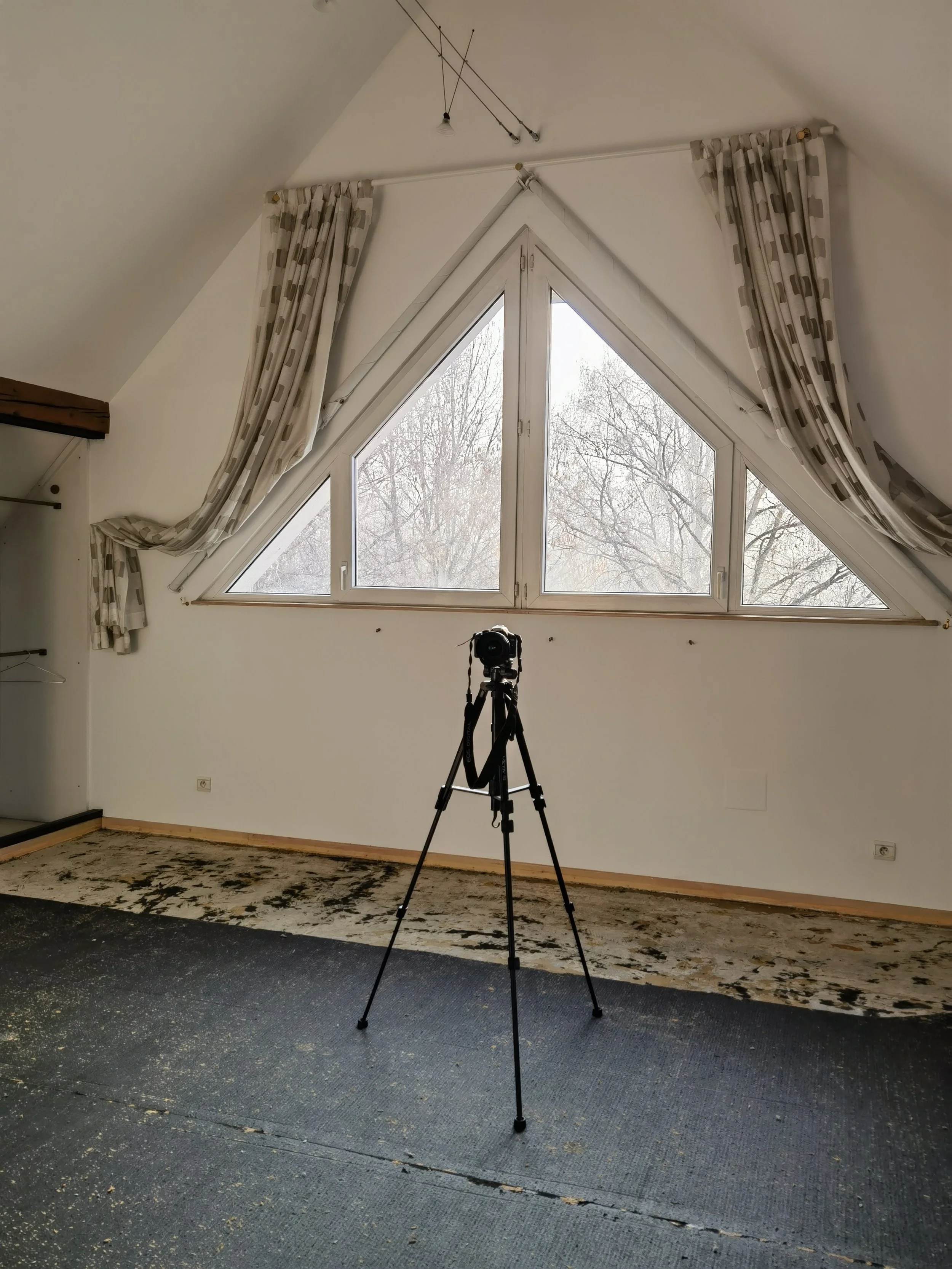 Une pièce avec une grande fenêtre en forme de triangle, des rideaux à carreaux, un tapis et un appareil photo sur un trépied.