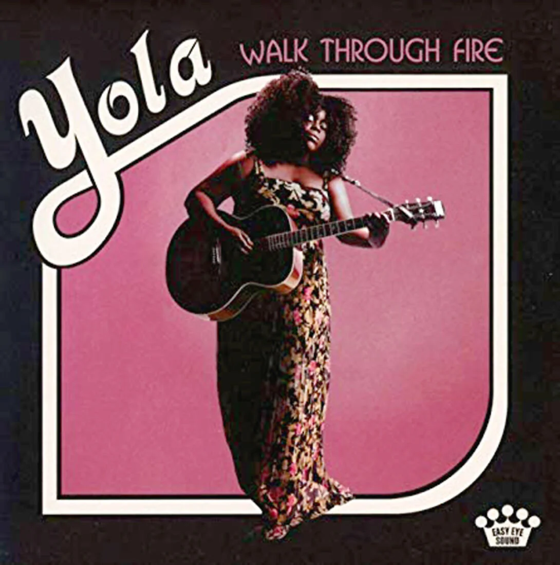 Image of a women playing a guitar in a floral dress infront of a pink backdrop with top text "Yola" "Walk through Fire"