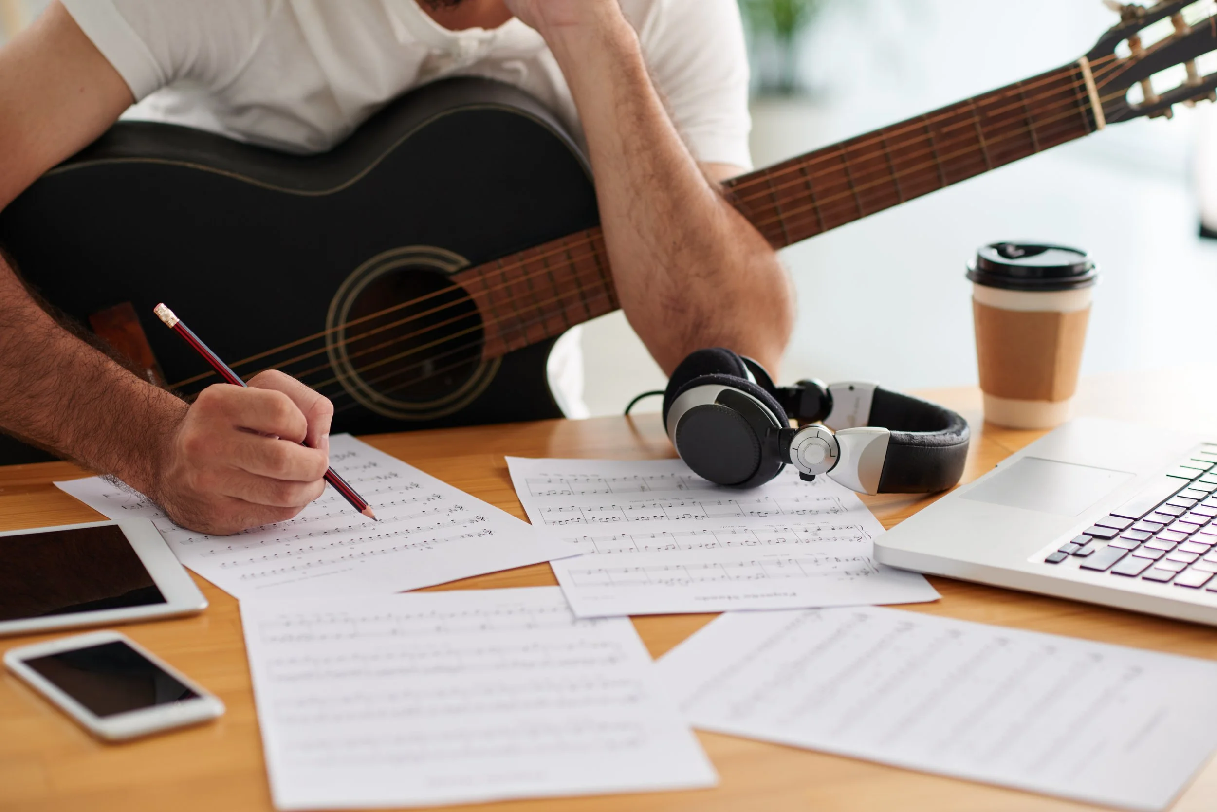 Image of a guy sitting with a guitar at a table writing on a music sheet with a laptop and headphones to his left