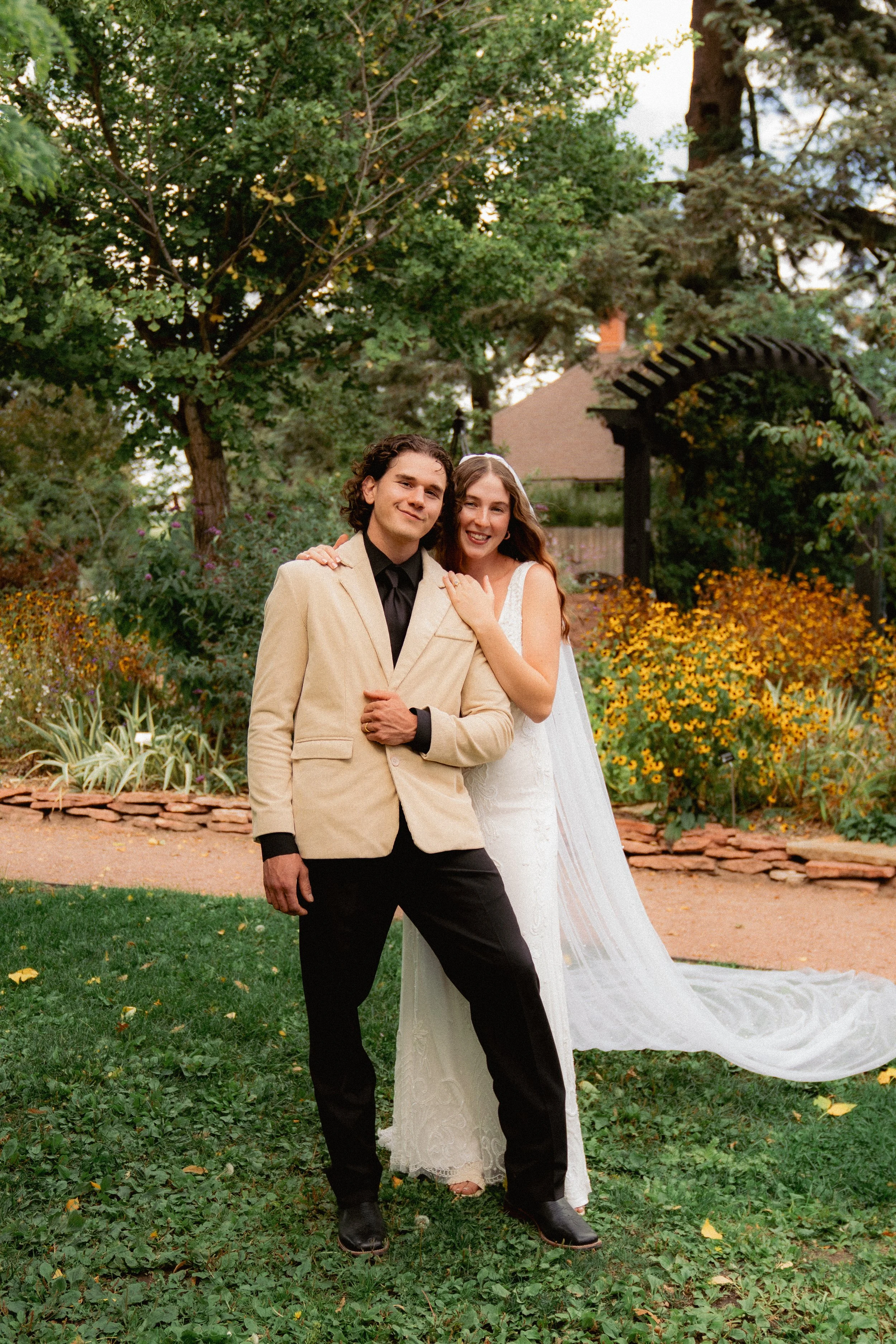 A couple dressed in wedding attire standing close together in a garden with trees, bushes, and yellow flowers in the background.