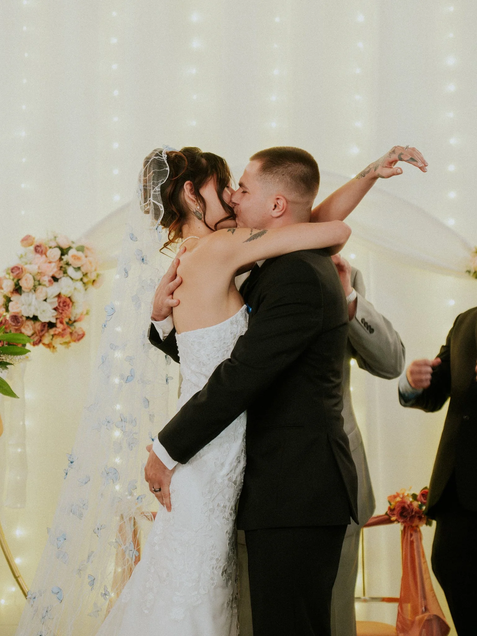 A bride and groom share a kiss during their wedding ceremony, with the bride wearing a white lace dress and veil, and the groom dressed in a black suit, in a decorated venue with floral arrangements and fairy lights.