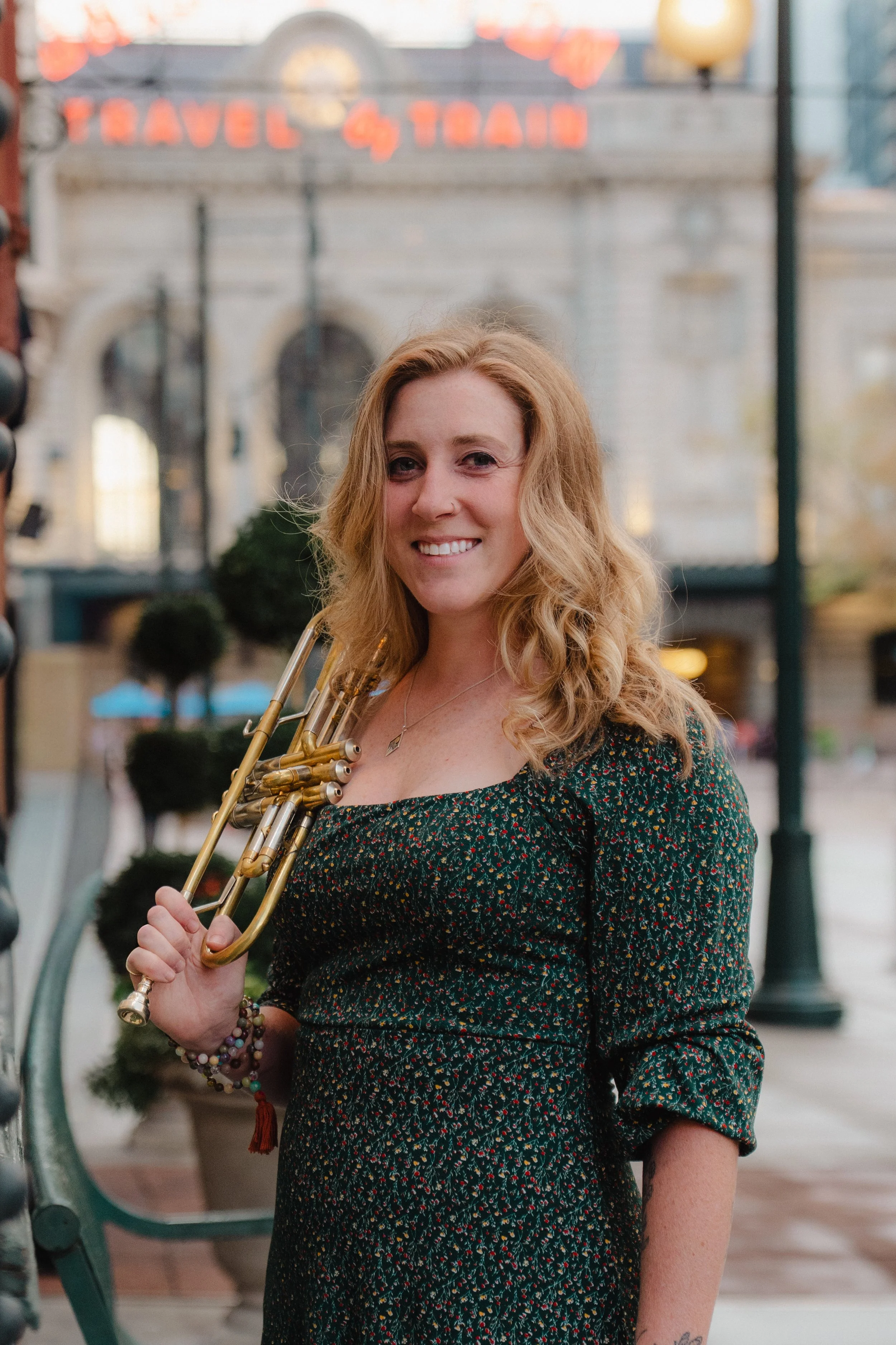 A woman with curly blonde hair holding a trumpet outdoors in a city setting, smiling at the camera.