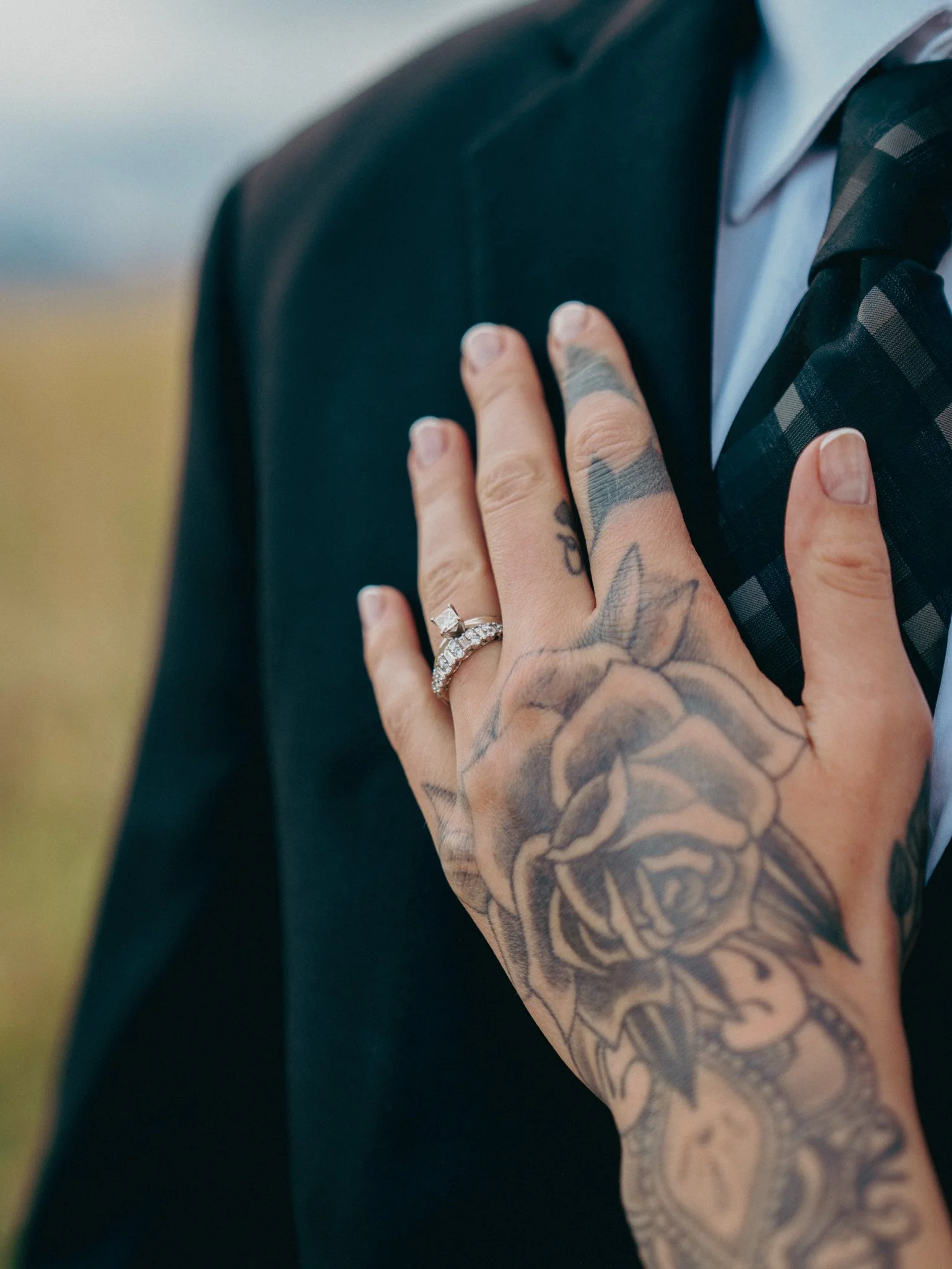 Close-up of a person's hand with tattoos, wearing a silver wedding and engagement ring, resting on the chest of a man in a suit and tie.