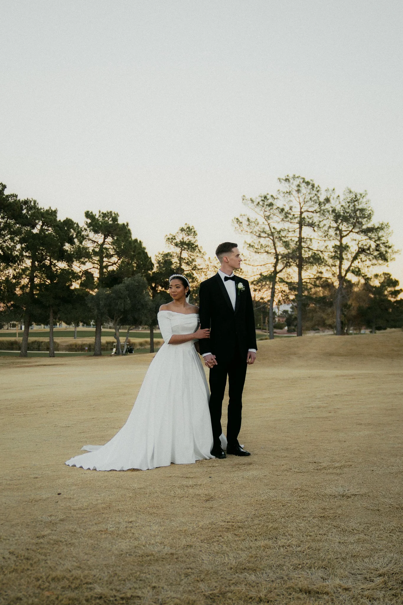 A bride in a white off-shoulder wedding gown holding hands with a groom in a black tuxedo, standing on a grassy field with trees and a sunset sky in the background.