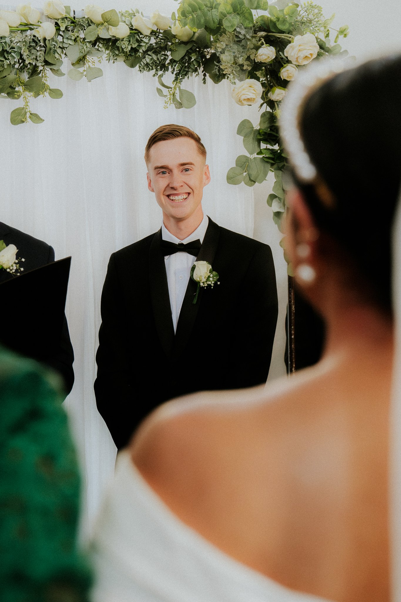 A groom in a tuxedo standing under a floral arch during a wedding ceremony, smiling at the bride.