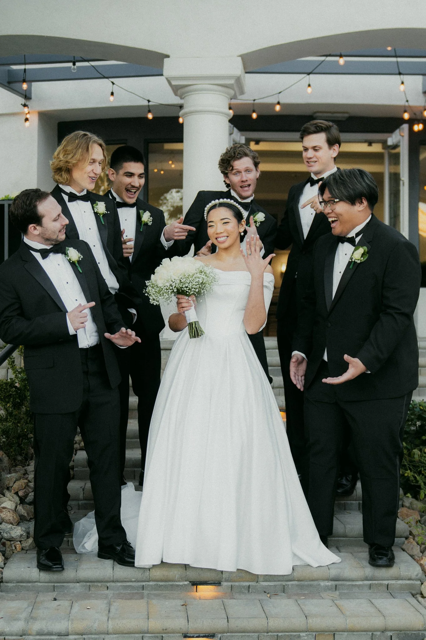 A bride in a white wedding dress with a bouquet and seven groomsmen in black tuxedos standing on steps outside a building, celebrating at a wedding.