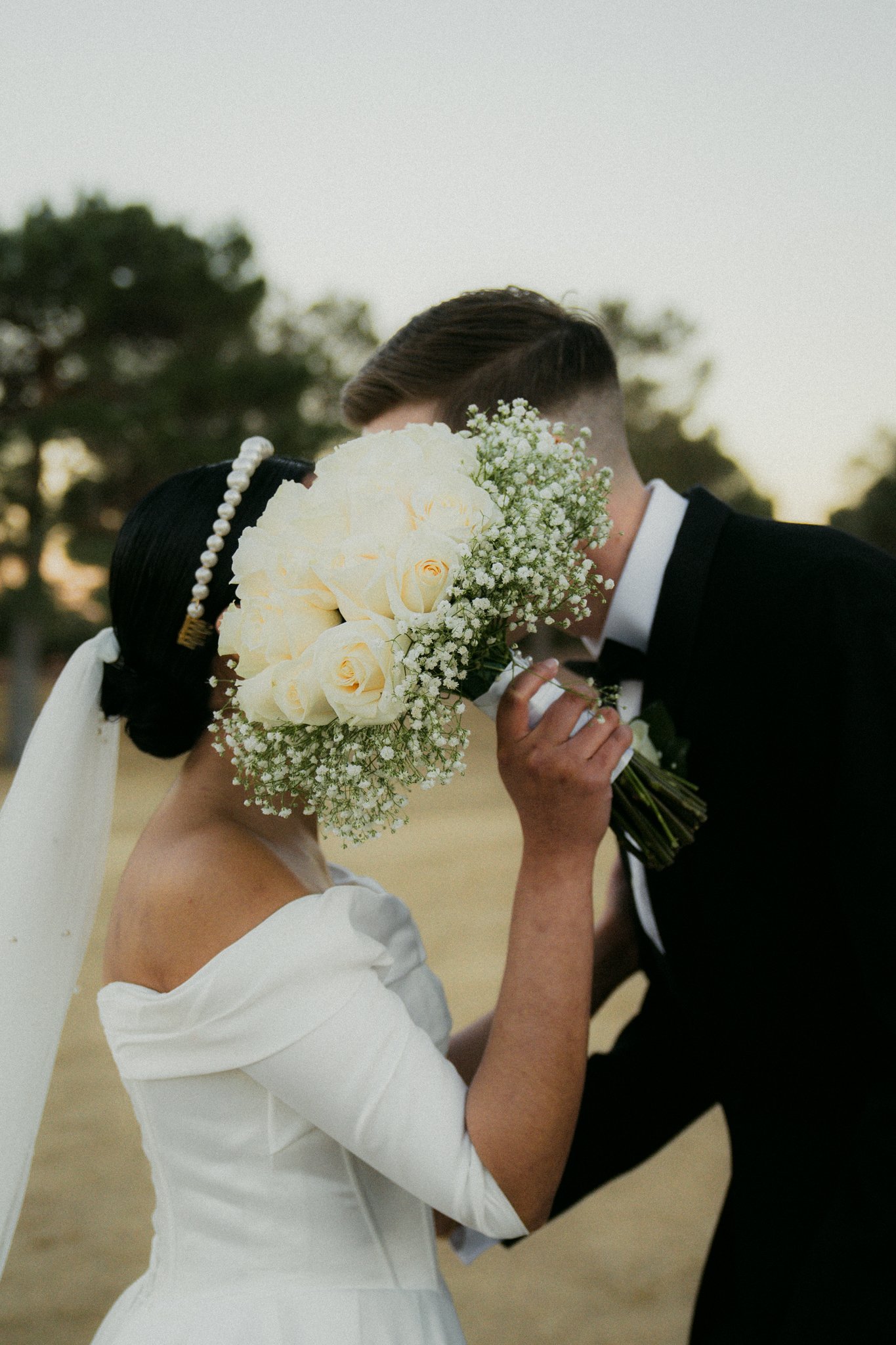 A bride and groom share a kiss, with the bride holding a bouquet of white roses and baby's breath, partially covering their faces. The bride wears a white off-shoulder dress and a pearl headband, while the groom is dressed in a black tuxedo with a wh
