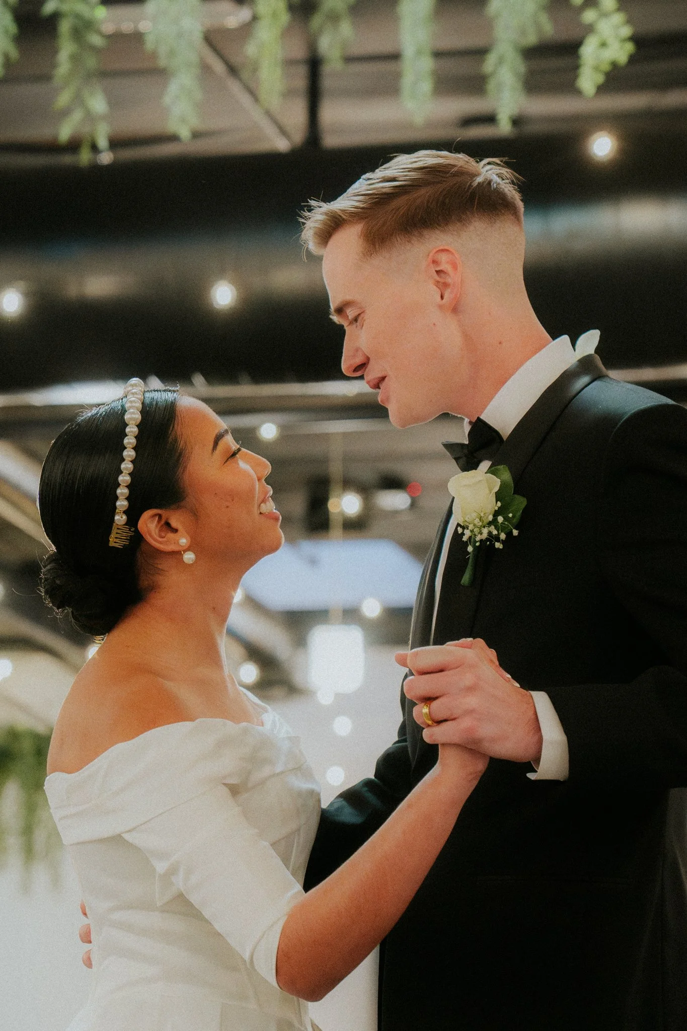 A bride and groom dance closely during their wedding reception, gazing into each other's eyes, indoors with hanging green plants overhead.