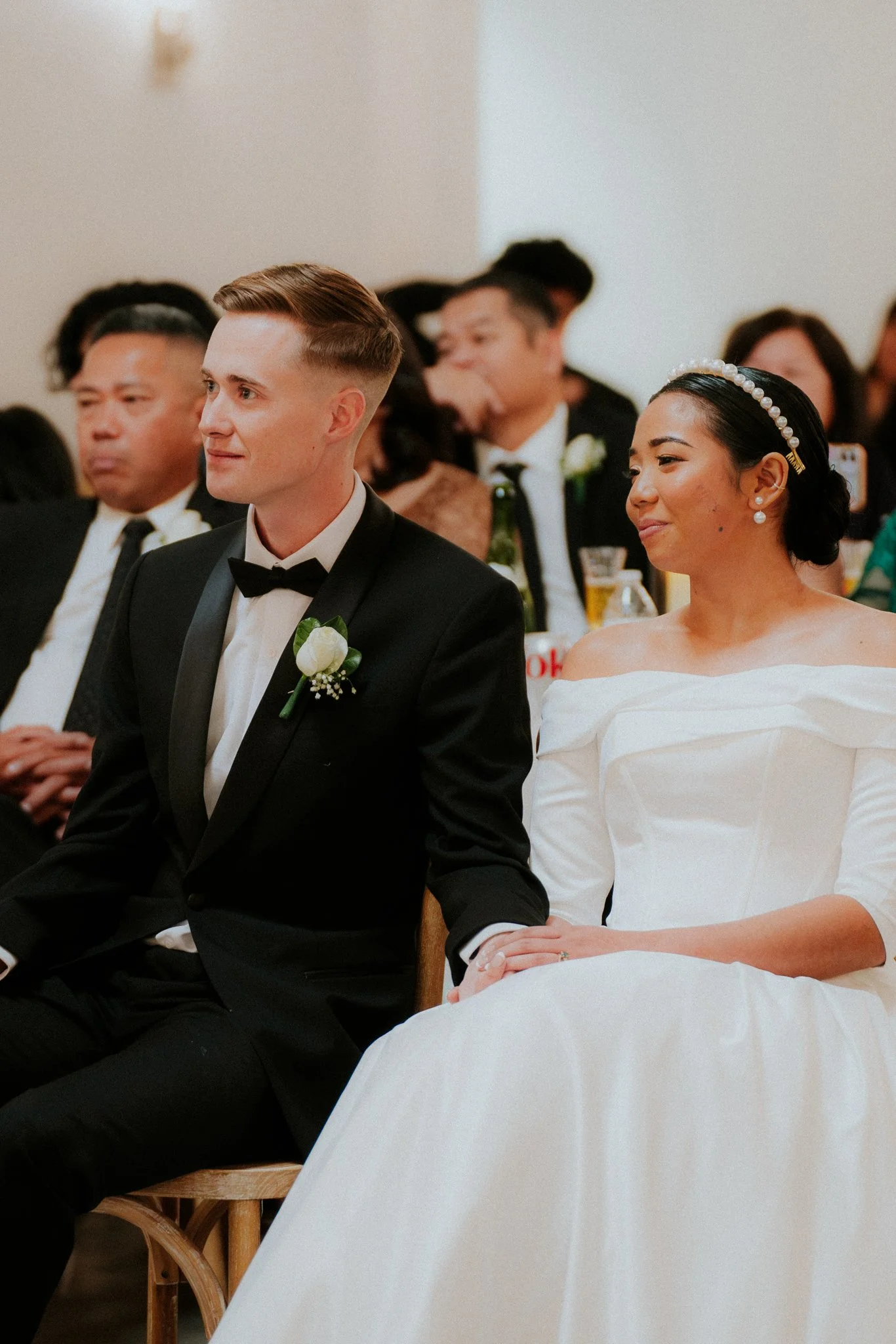 A bride and groom sitting together during their wedding ceremony, holding hands and smiling, with guests in the background.