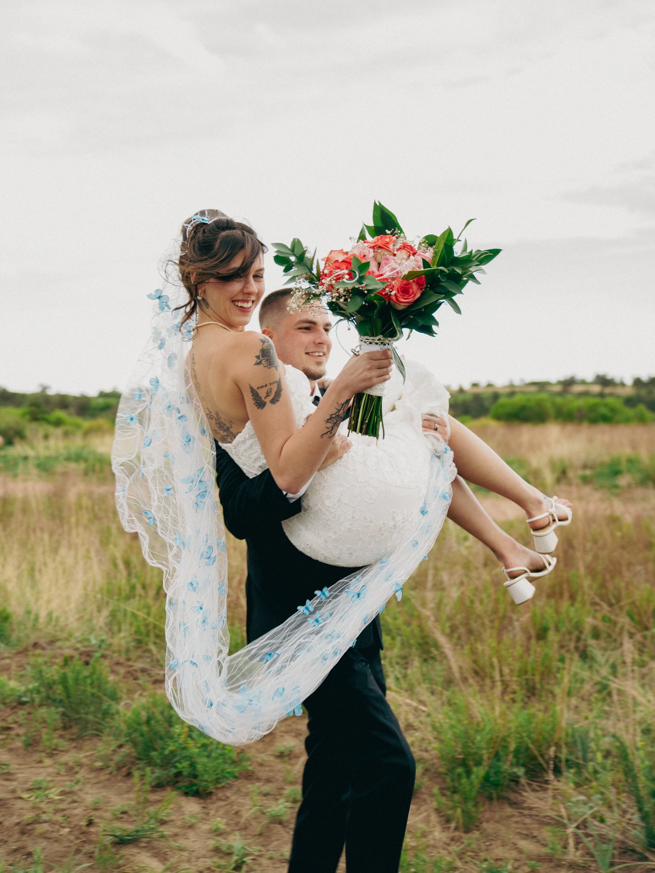 A man in a suit holding a woman in a wedding dress with a bouquet of flowers in a grassy field.