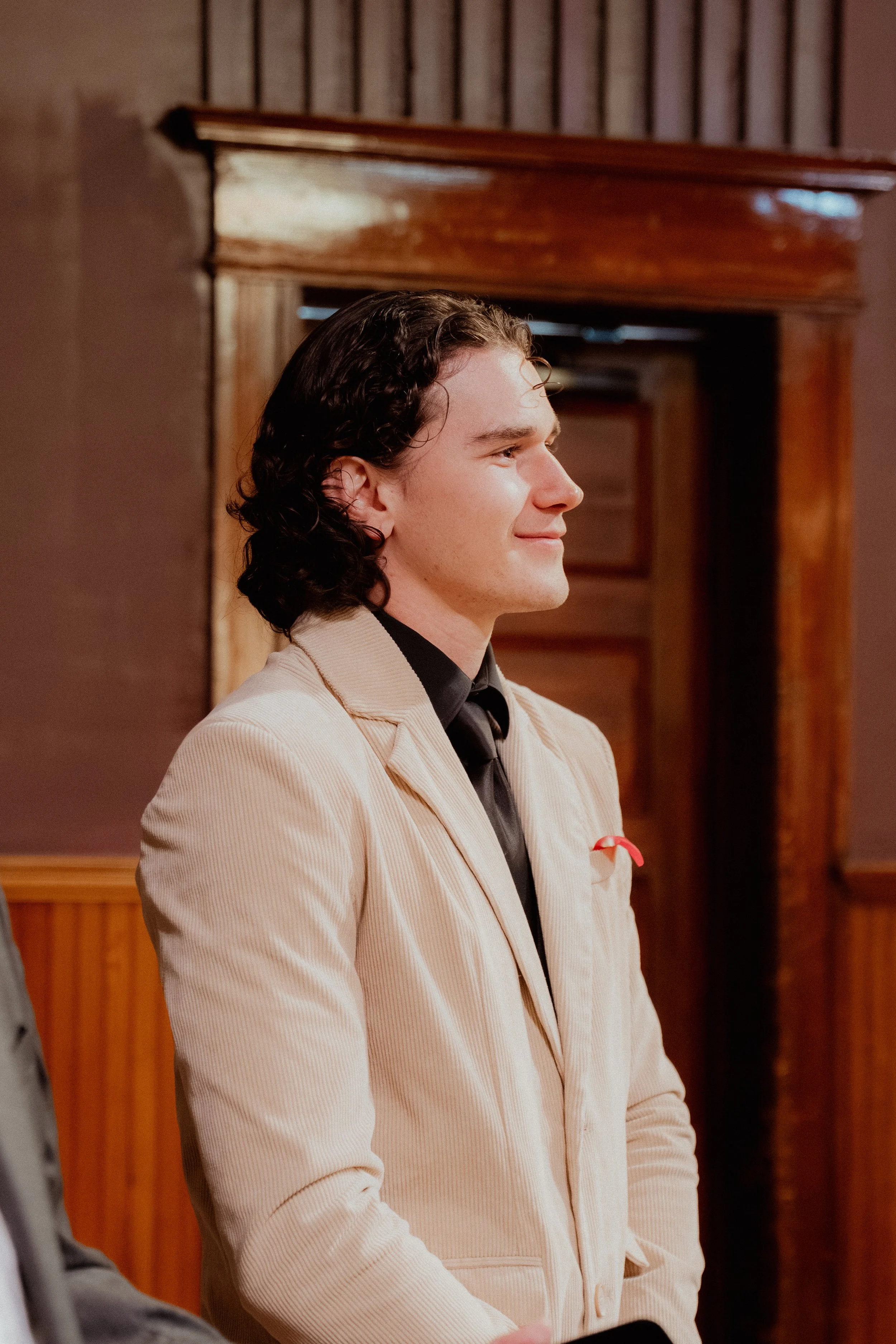 Side profile of a young man with dark, curly hair wearing a beige blazer over a black shirt, smiling softly in a warm-toned room with wooden decor.
