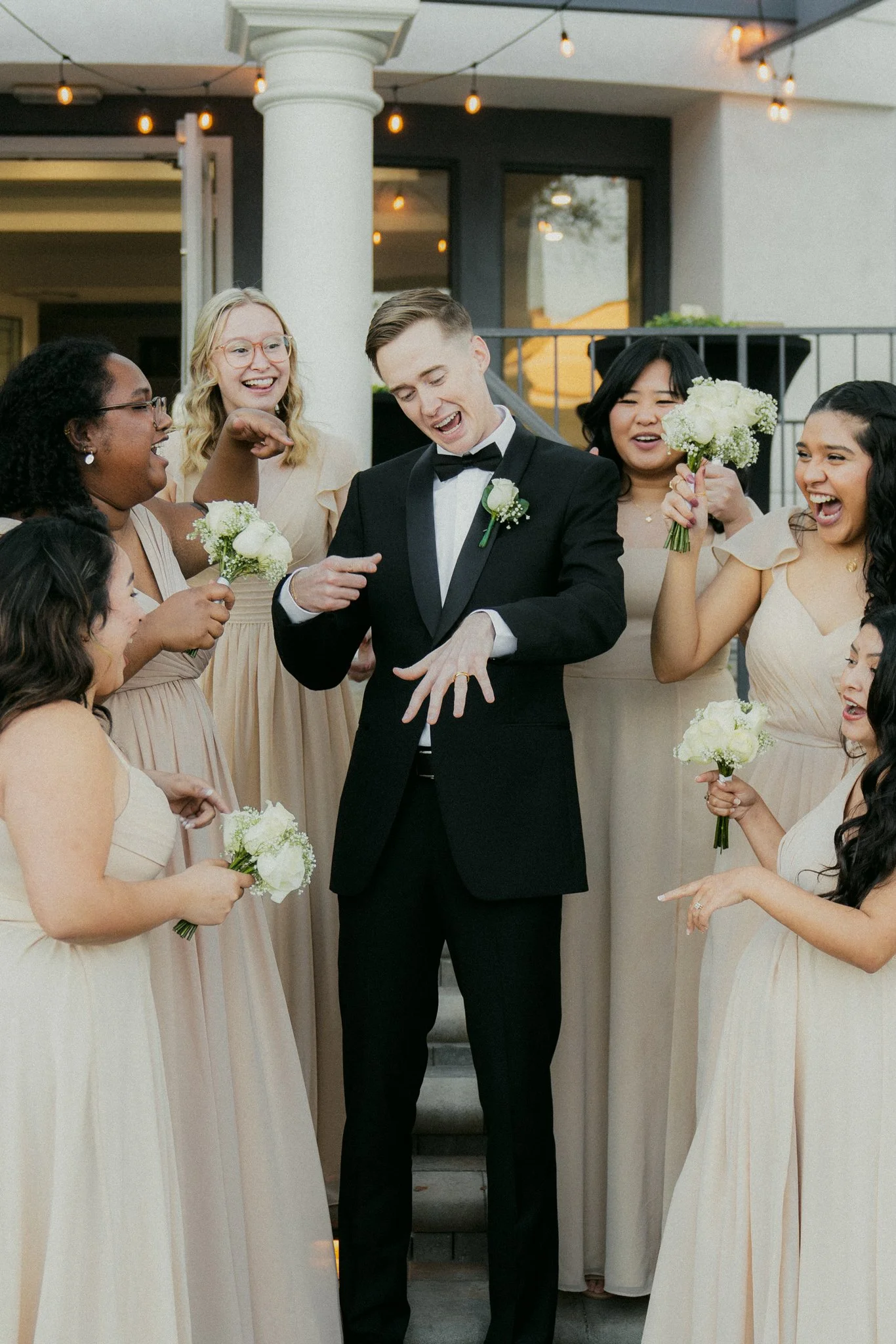 A group of women dressed in beige dresses and a man in a black tuxedo celebrating a wedding, with the man showing off a wedding ring on his finger.