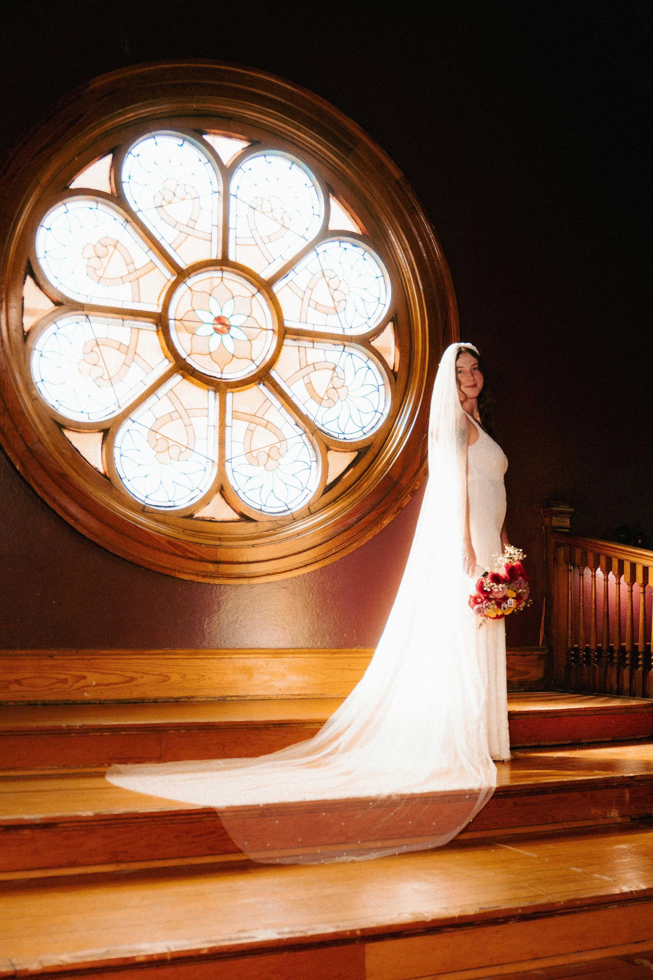 Bride in wedding dress holding bouquet standing near a large circular stained glass window with wooden frame, inside a wooden interior.