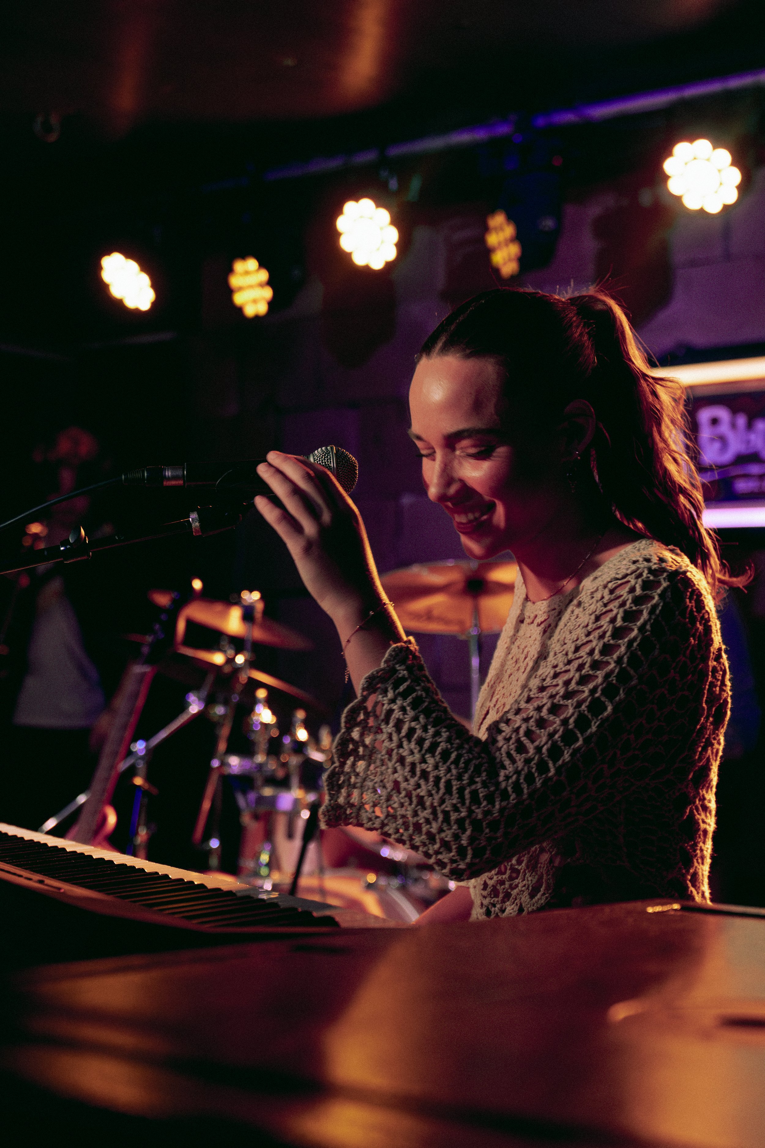 A woman smiling and singing into a microphone while playing the keyboard at a dimly lit live music venue.
