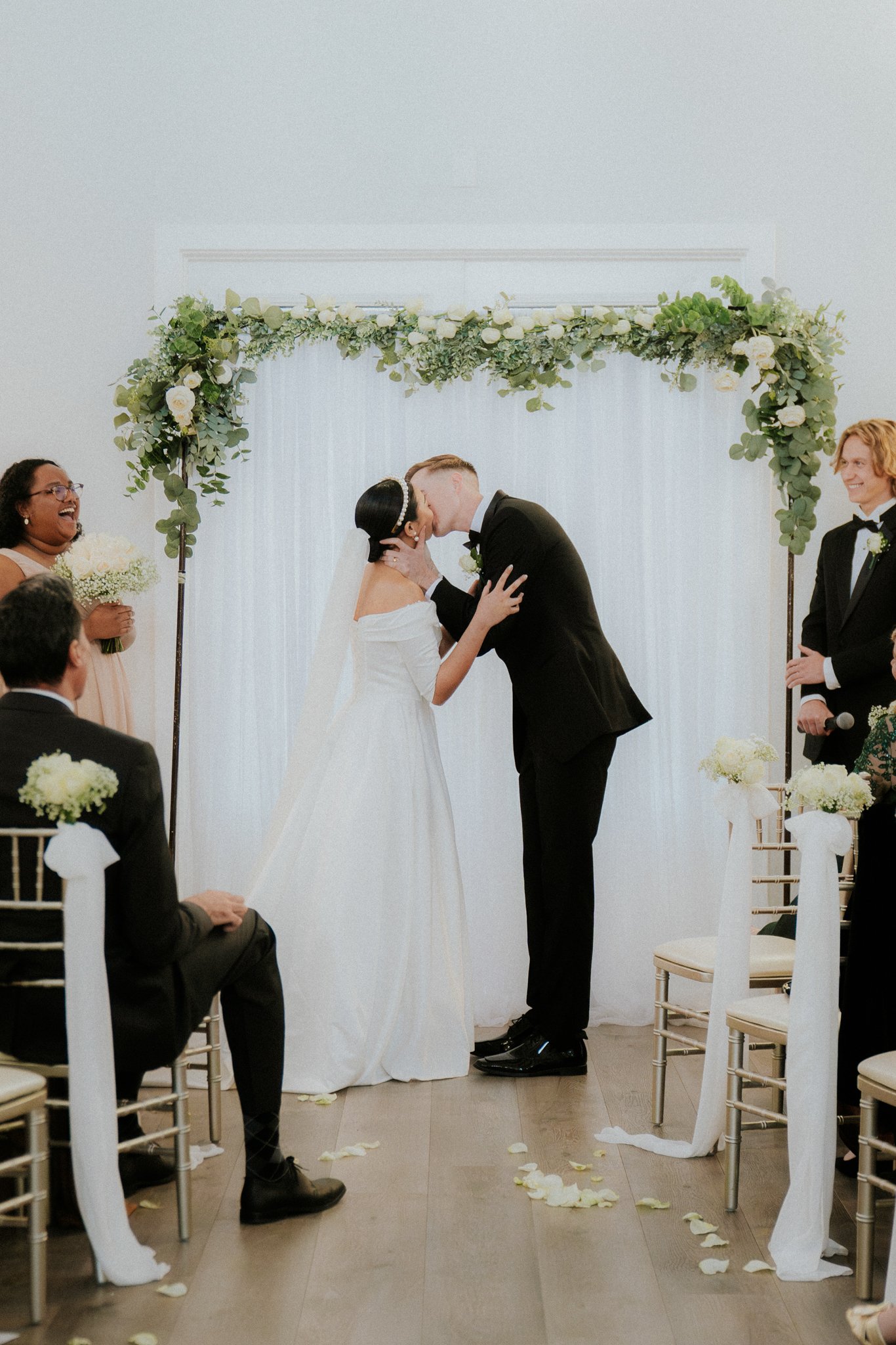 Couple sharing a kiss at their wedding ceremony, surrounded by joyful guests, with floral decorations and white fabric backdrop.