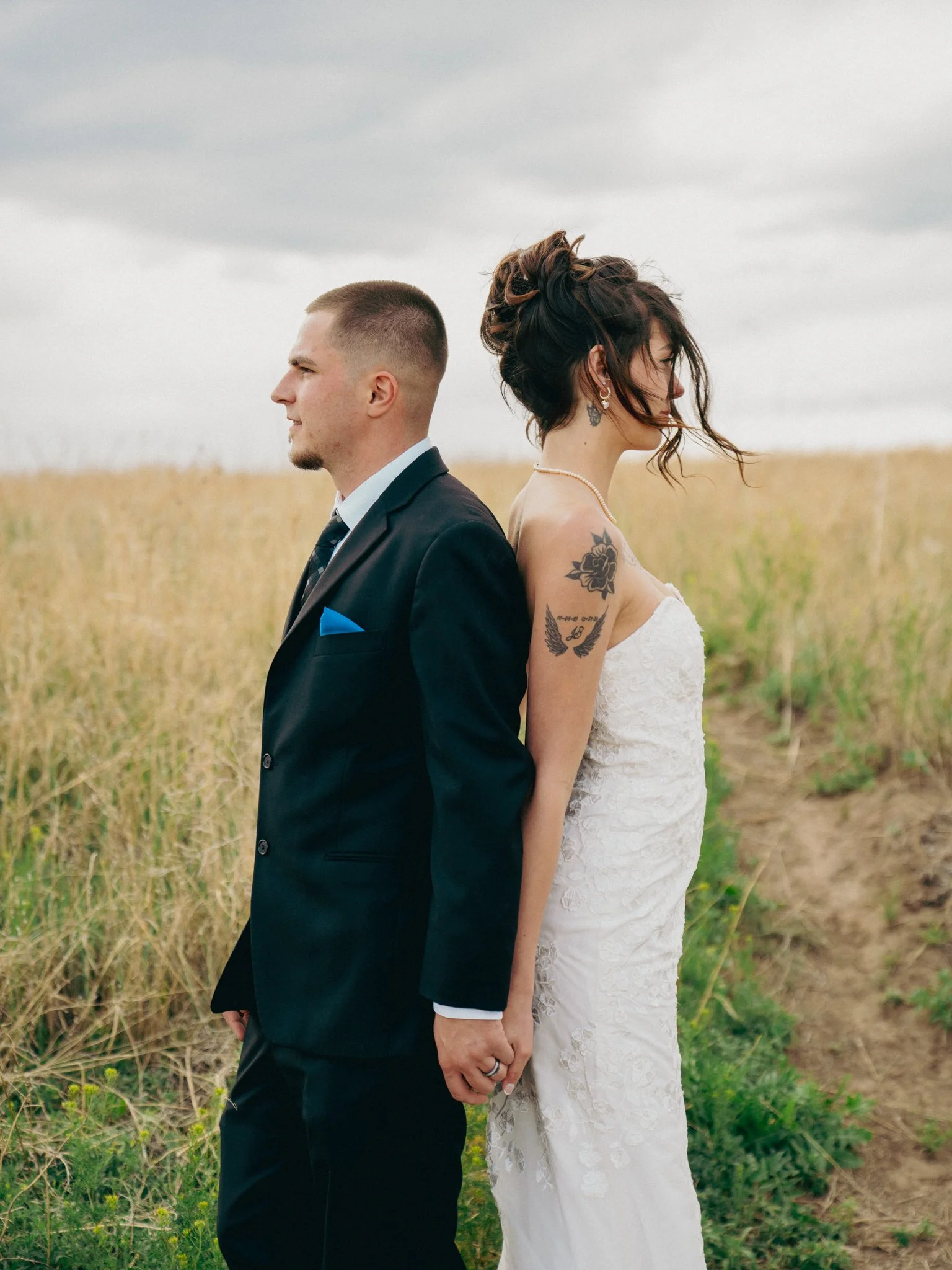 A bride and groom stand back to back in a field with tall grass, holding hands with fingers intertwined, during an outdoor wedding photoshoot. The groom is wearing a black suit with a white shirt and a blue pocket square. The bride is wearing a strap
