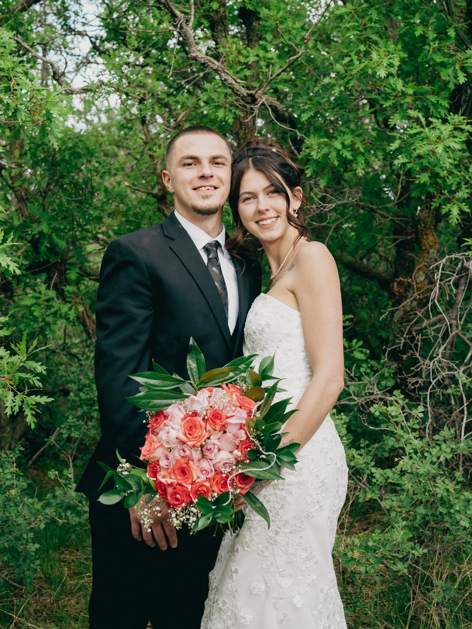 A newlywed couple standing outdoors in front of green trees, with the groom in a black suit and tie, and the bride in a white lace wedding dress, holding a bouquet of pink and orange roses.