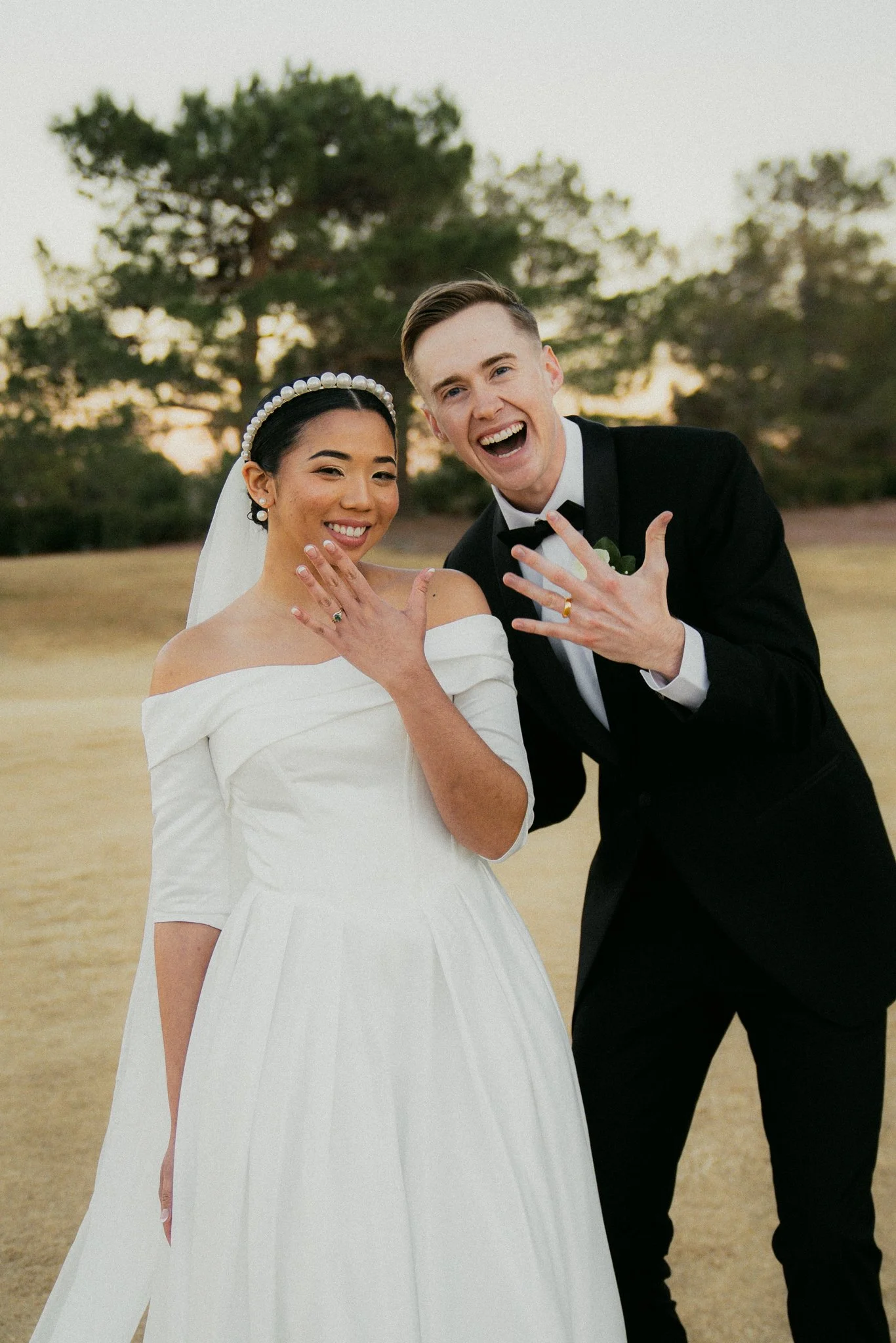 A newlywed couple celebrating outdoors, showing off their wedding rings with big smiles. The bride is wearing a white off-the-shoulder wedding gown and veil, and the groom is in a black tuxedo with a bow tie, standing in front of a backdrop of trees 
