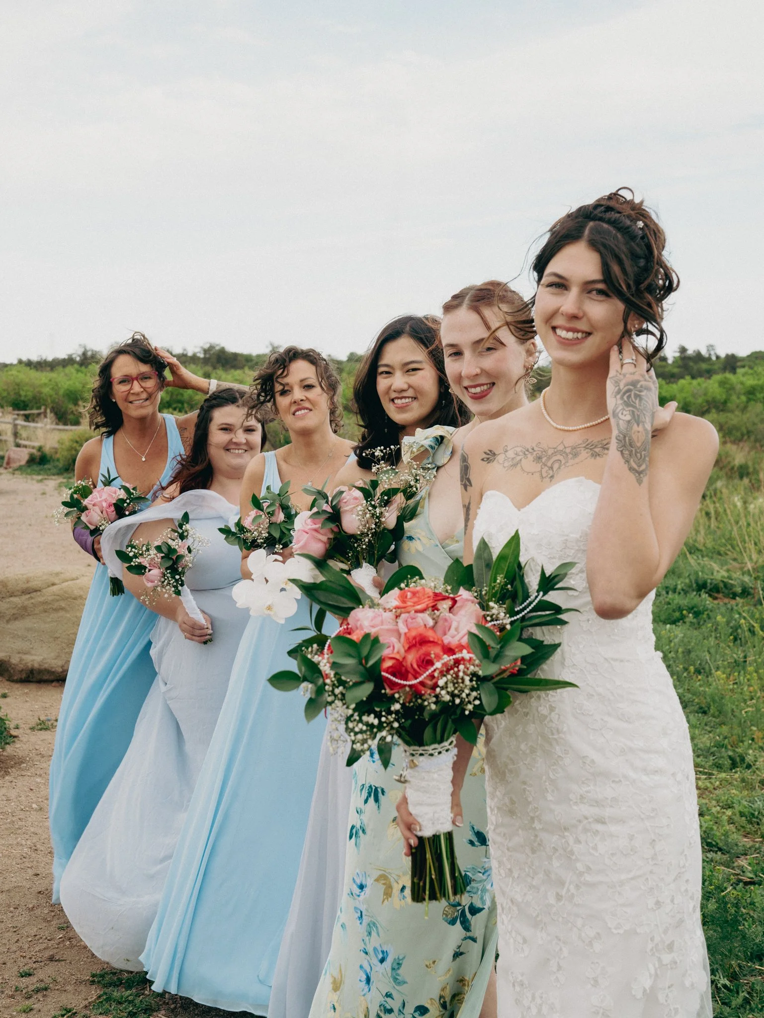 A wedding party of six women, standing outdoors on a grassy area with a cloudy sky. The bride, dressed in a white lace gown, is at the front holding a bouquet of pink and red flowers. The bridesmaids behind her are in pastel blue and floral dresses, 