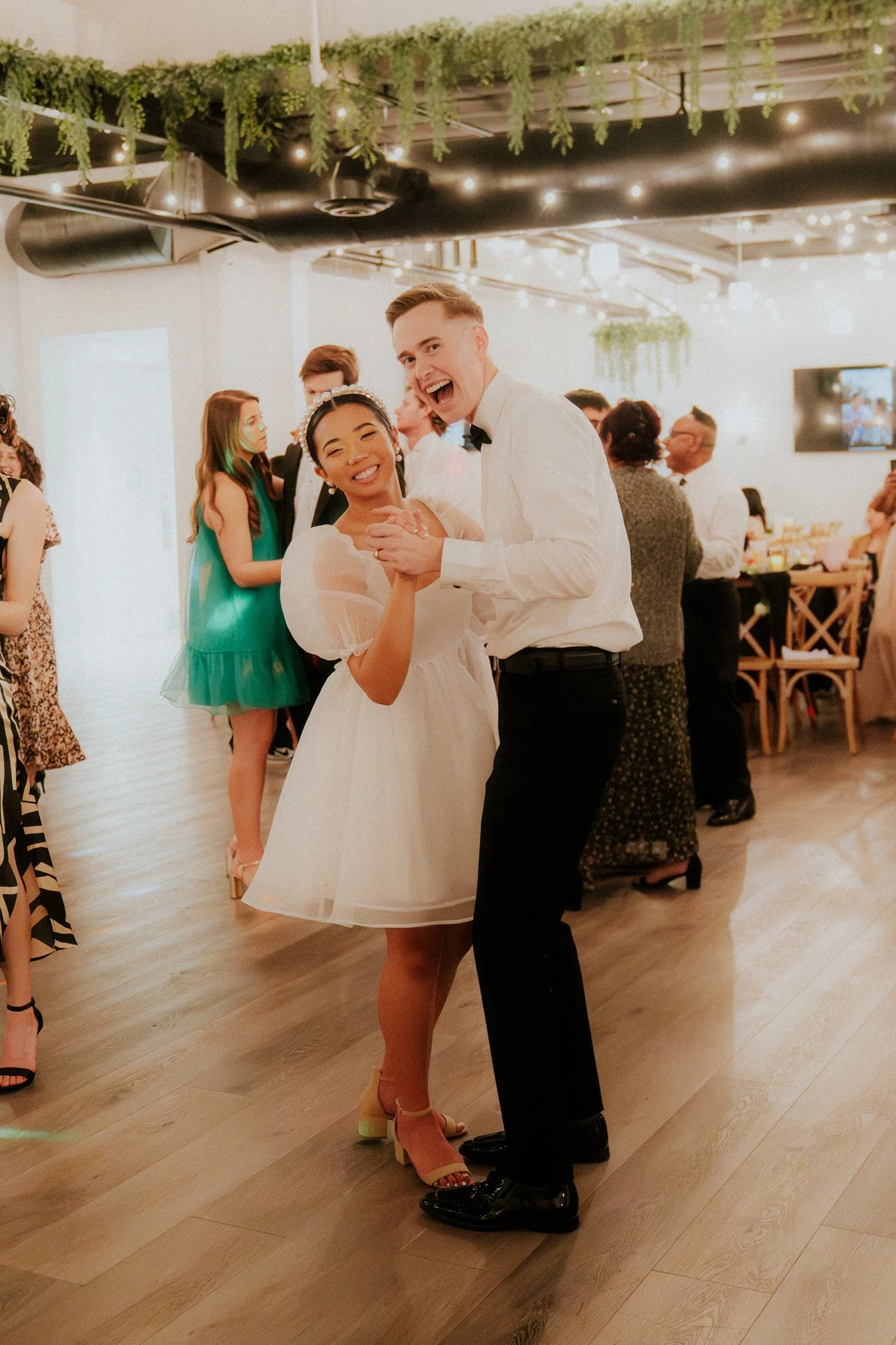 A young couple dancing at a celebration, with other guests in the background, in a warmly lit venue decorated with greenery and string lights.
