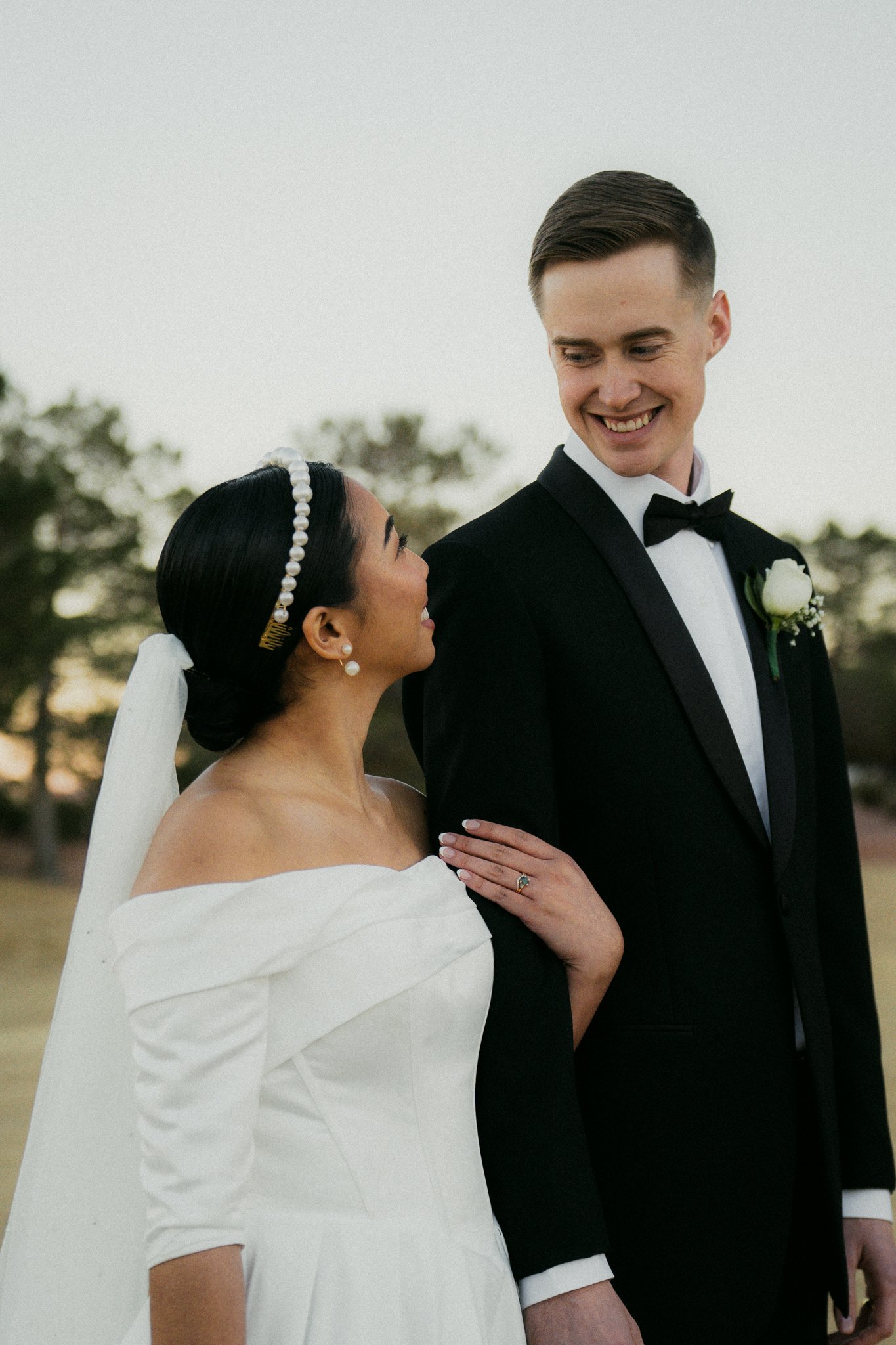 A bride and groom on their wedding day outdoors, smiling at each other. The groom wears a black tuxedo with a bow tie and boutonniere, and the bride wears a white off-shoulder gown with a pearl headband and veil.