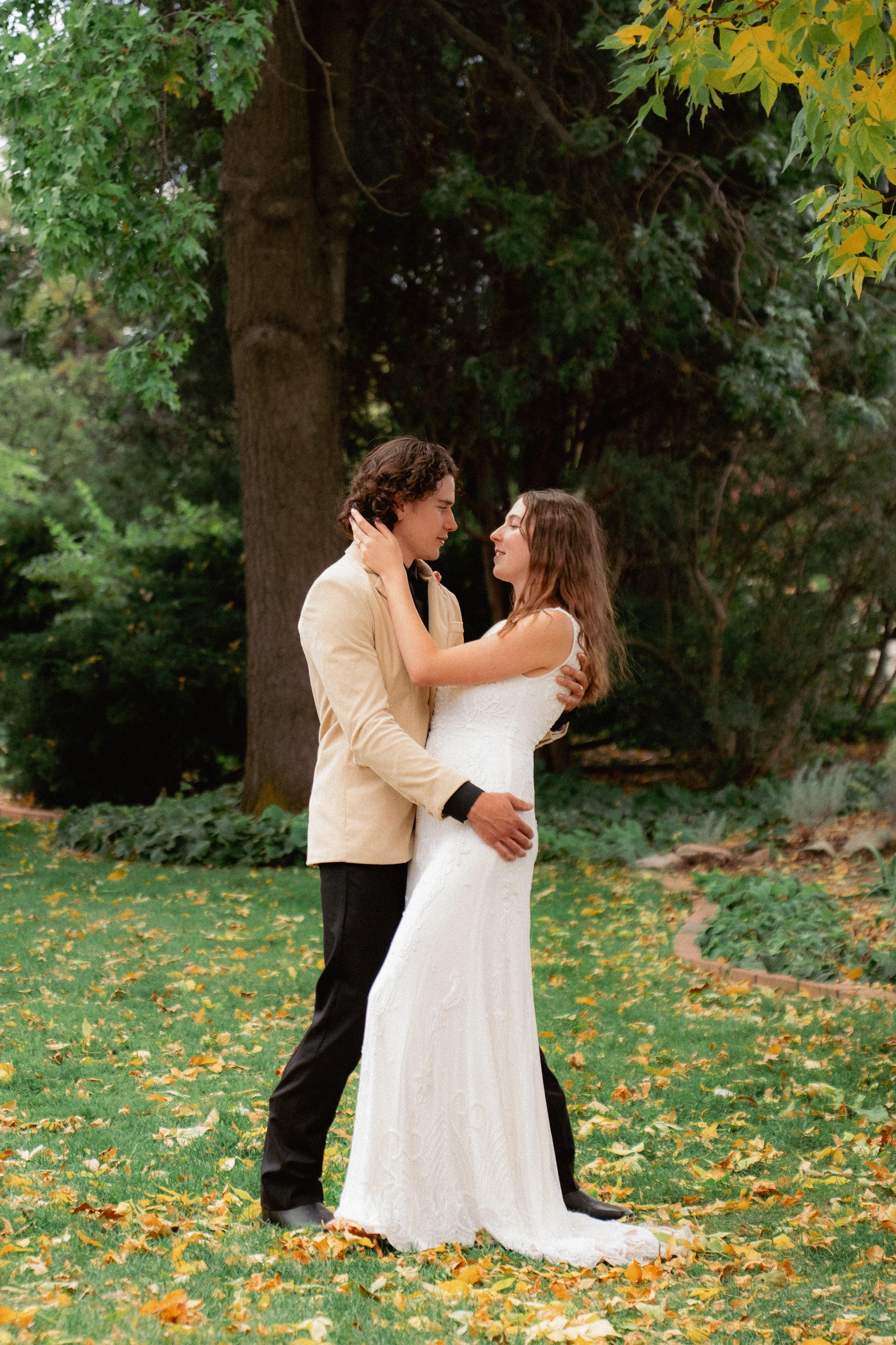 A couple in wedding attire sharing a romantic moment outdoors during fall, surrounded by trees with yellow leaves on the ground.