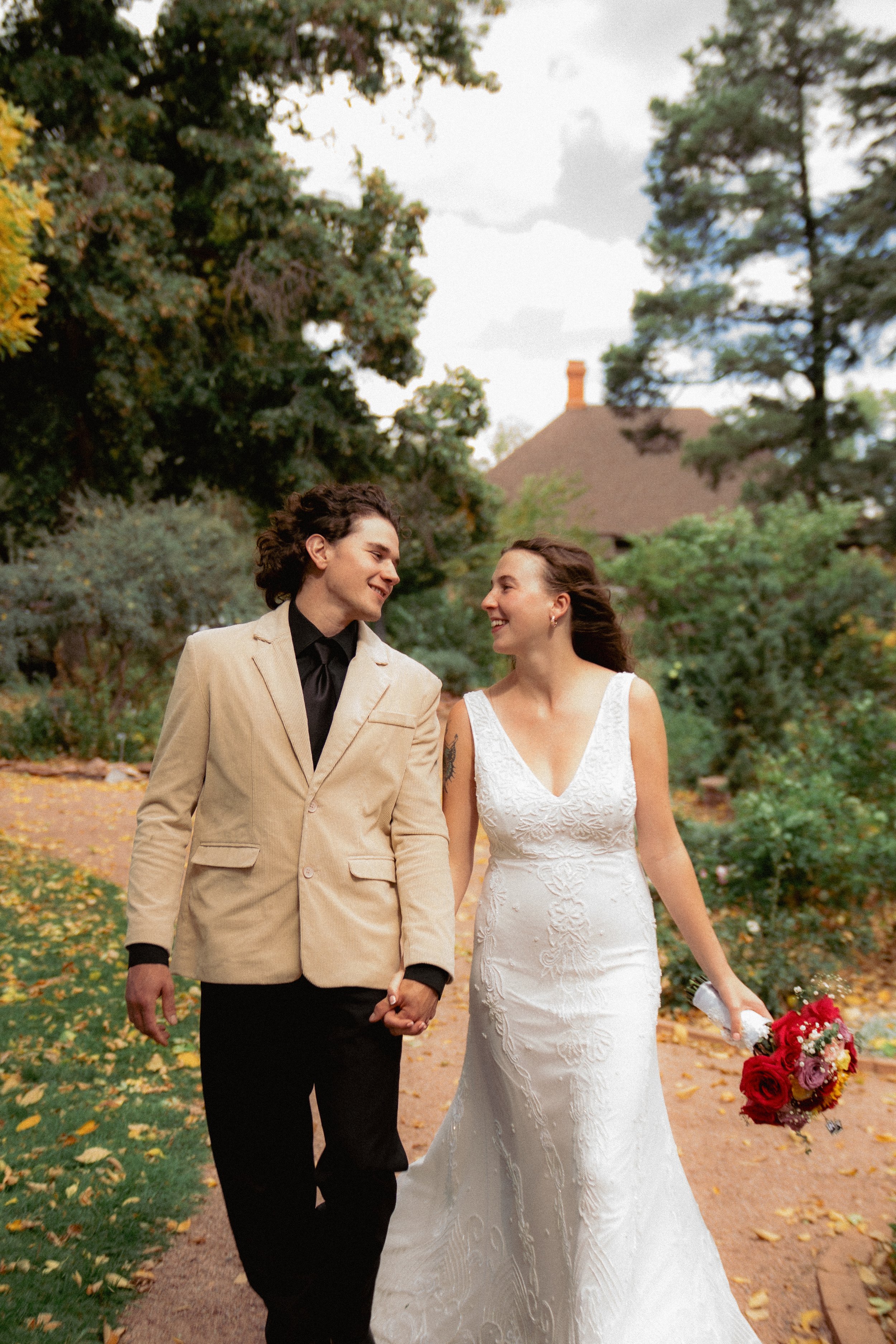A couple holding hands and walking in a park, dressed in wedding attire, smiling at each other. The woman is wearing a white wedding dress and holding a bouquet of red and pink flowers, while the man is wearing a beige blazer and black shirt. They ar
