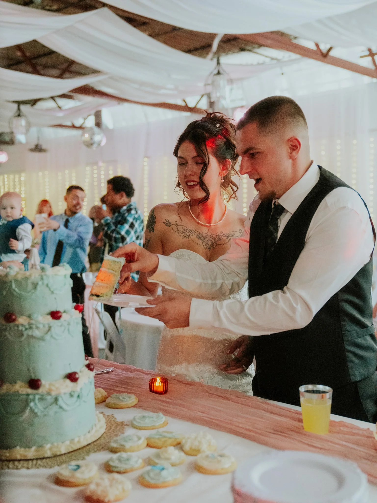 A bride and groom cutting and serving cake at their wedding reception. The cake is decorated with cherries and white icing, and there are cookies on the table.