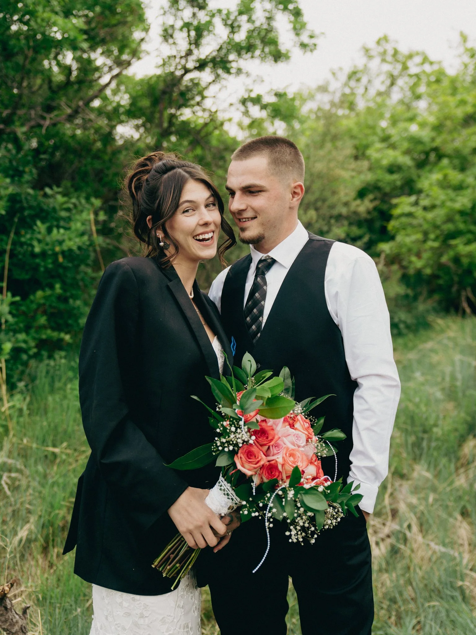 Bride and groom standing outdoors with greenery, the bride holding a bouquet of pink and peach roses, both smiling.