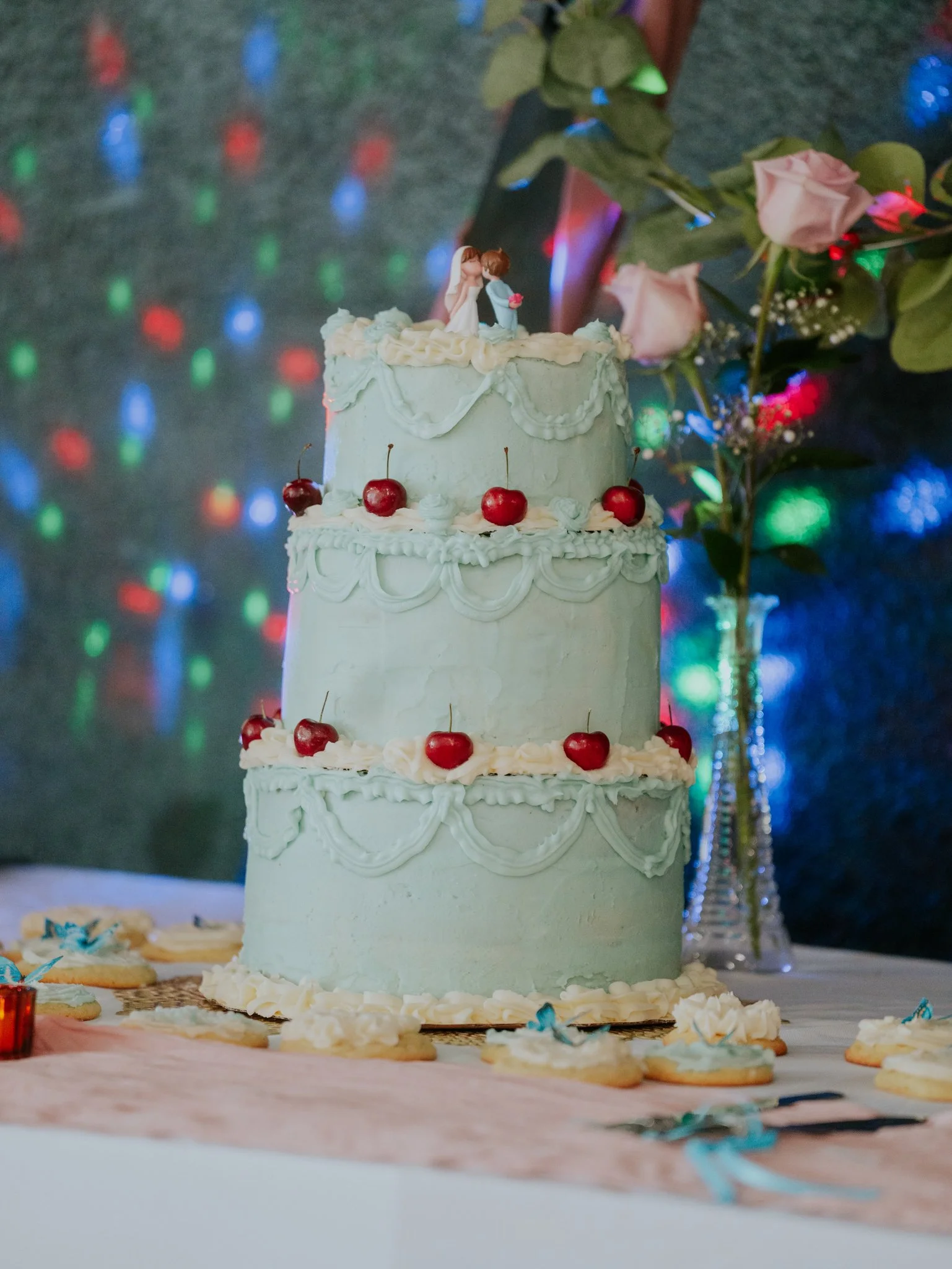 Three-tier wedding cake decorated with cherries and white icing, topped with a bride and groom figurine, and surrounded by cake cookies with blue bows, on a table with a pink cloth and a vase with pink roses and greenery, with multicolored lights in 