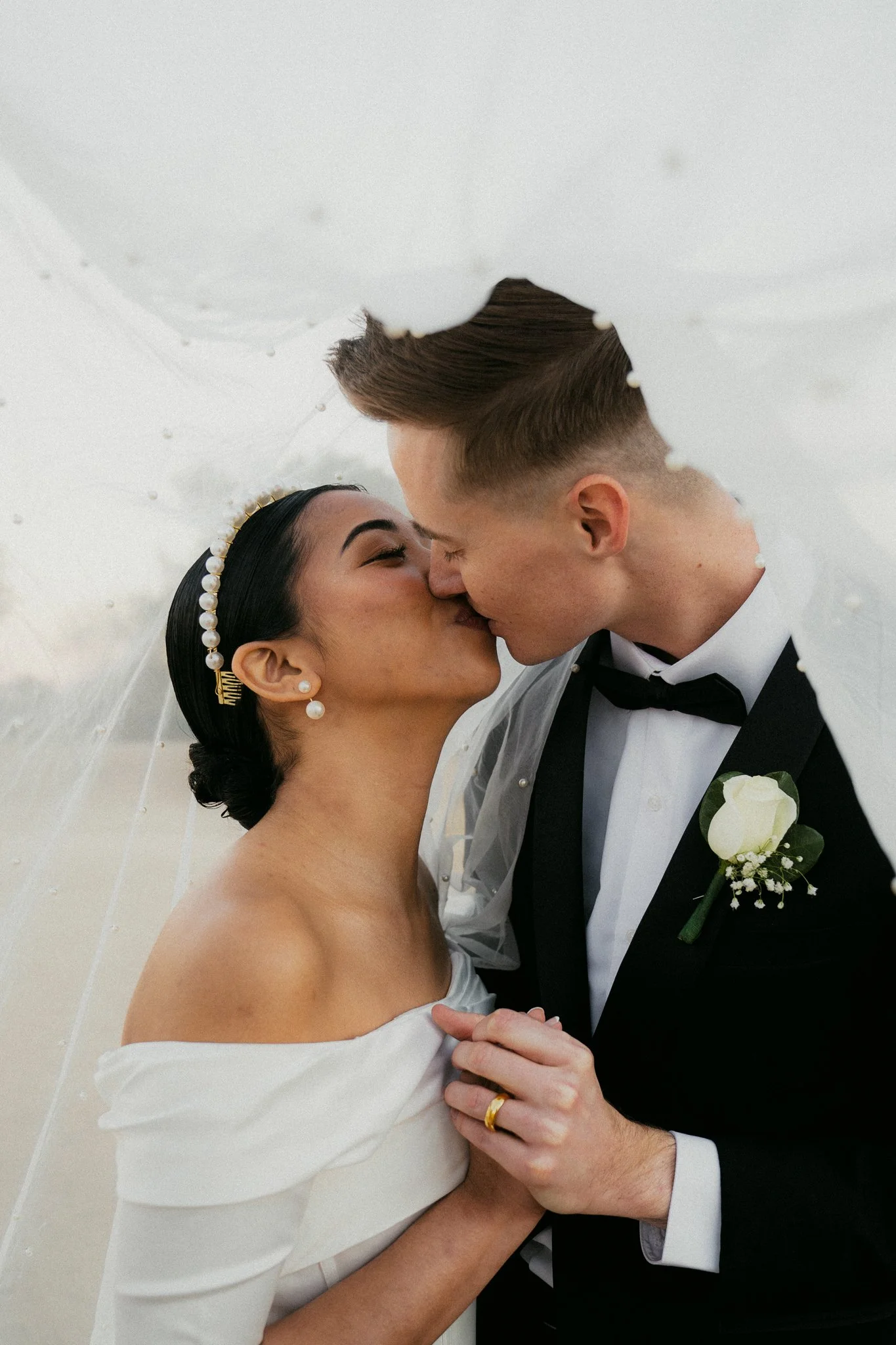 A newly married couple sharing a kiss, with the bride holding a transparent umbrella, in wedding attire.
