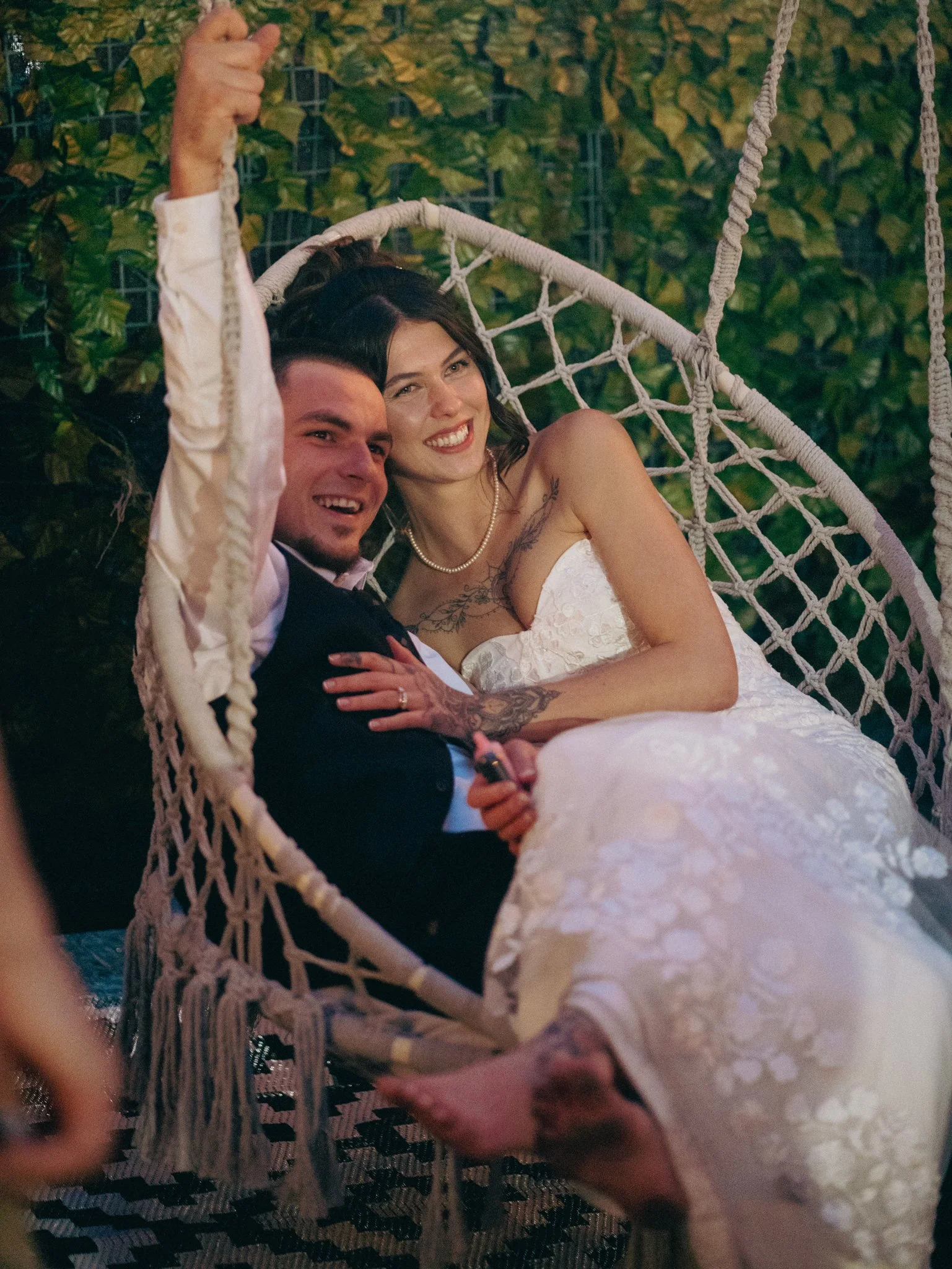 A newlywed couple sitting on a decorative hanging rope chair outdoors, smiling and celebrating their wedding day.