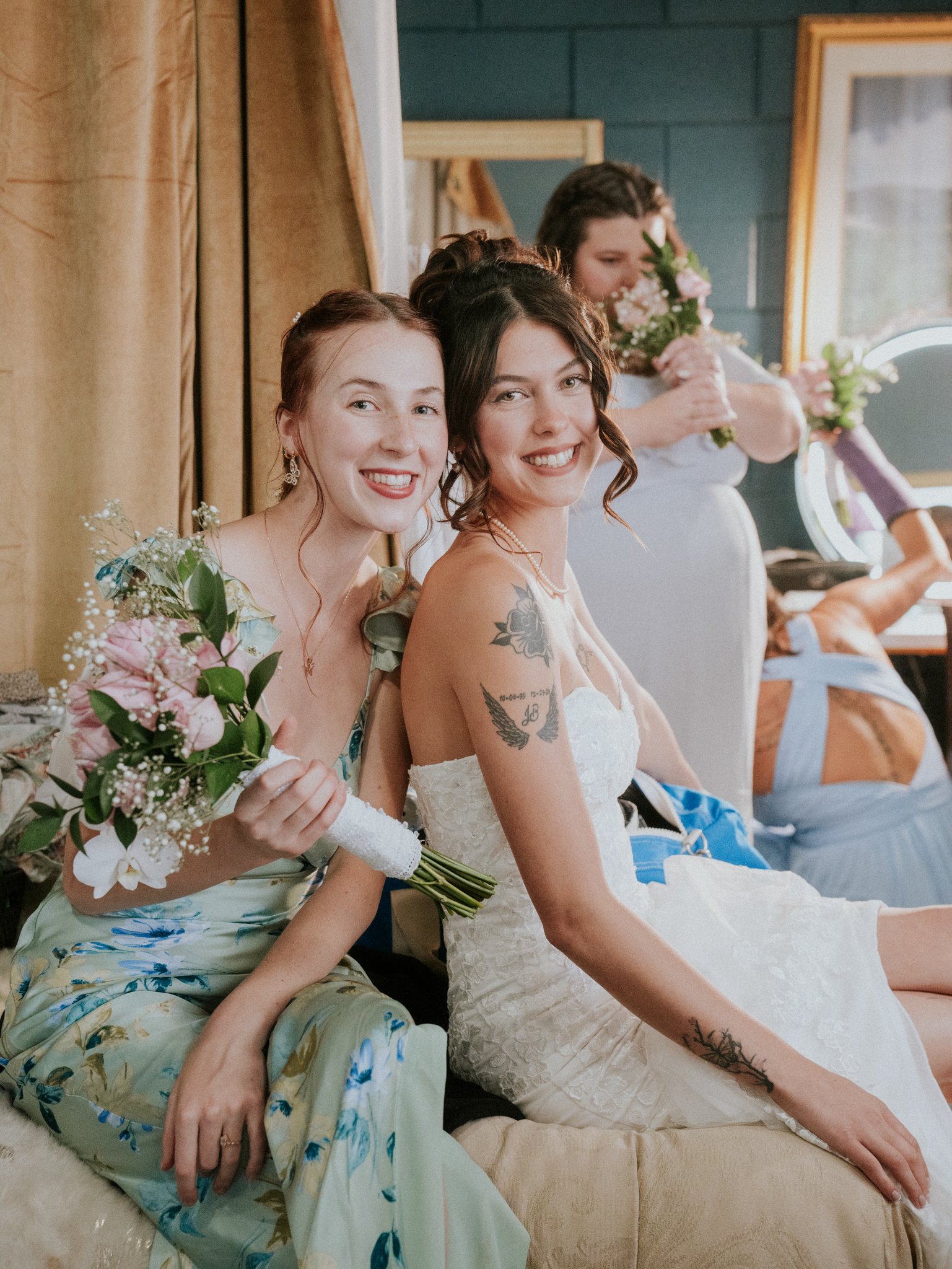 Two women smiling, one holding a bouquet of flowers, at a wedding reception. One woman is wearing a floral dress, the other a white wedding dress with tattoos on her left arm.