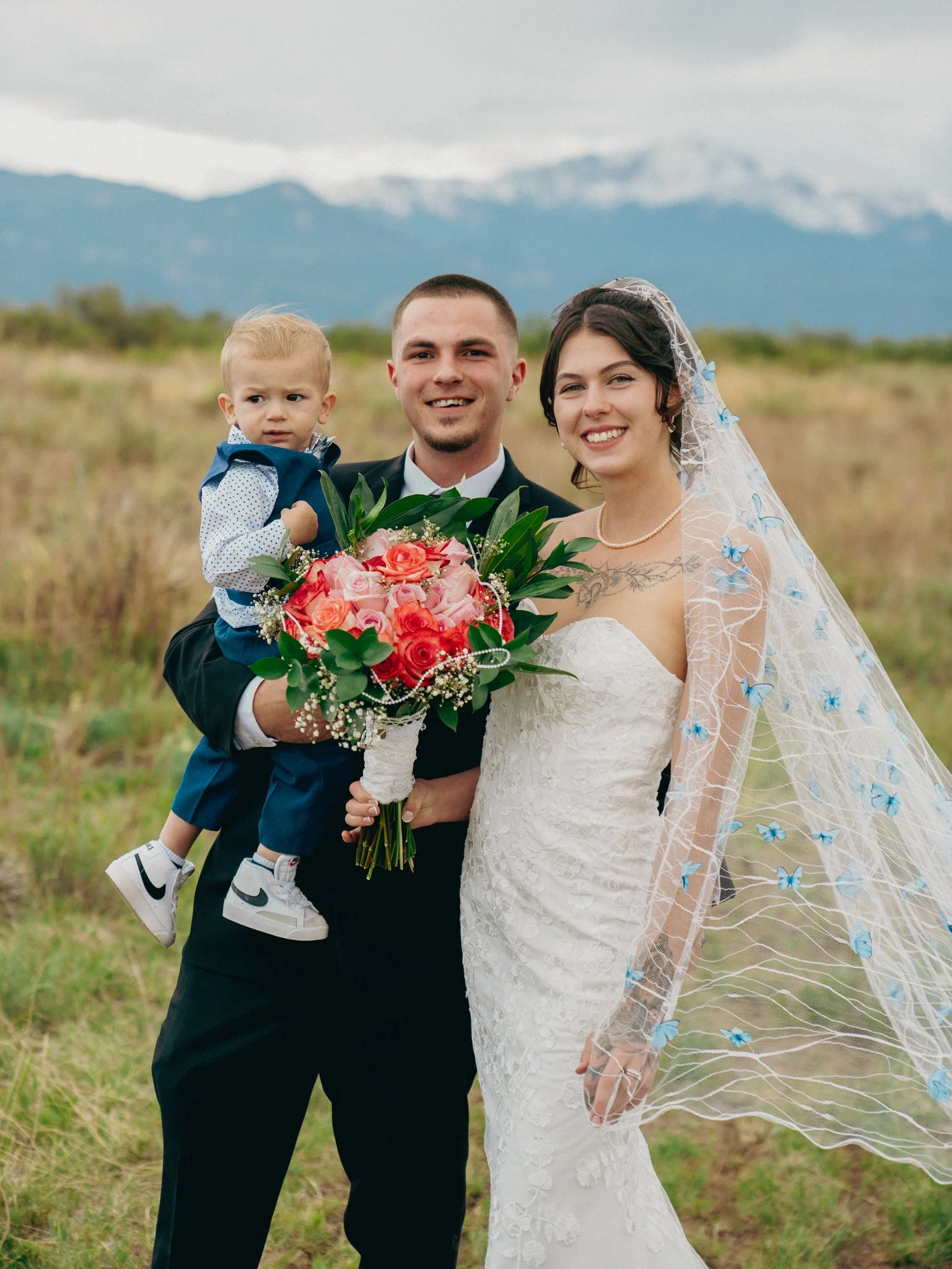 A newlywed couple with a young child outdoors in a field with mountains in the background, the groom holding a bouquet of pink and orange roses, the bride wearing a lace wedding dress with a veil decorated with blue butterflies.
