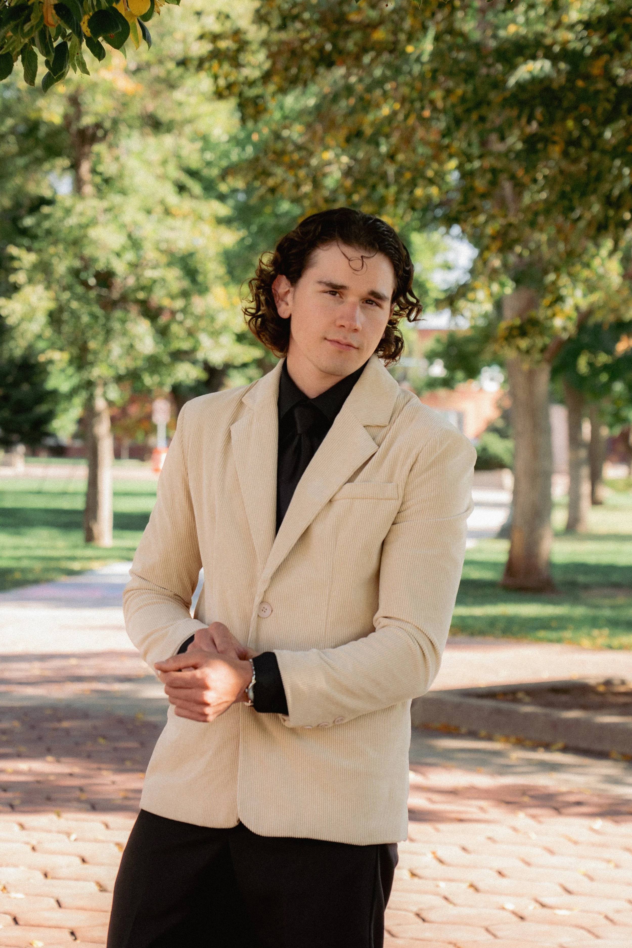 A young man with curly brown hair wearing a beige blazer over a black shirt, standing outdoors on a brick path with trees and greenery in the background.