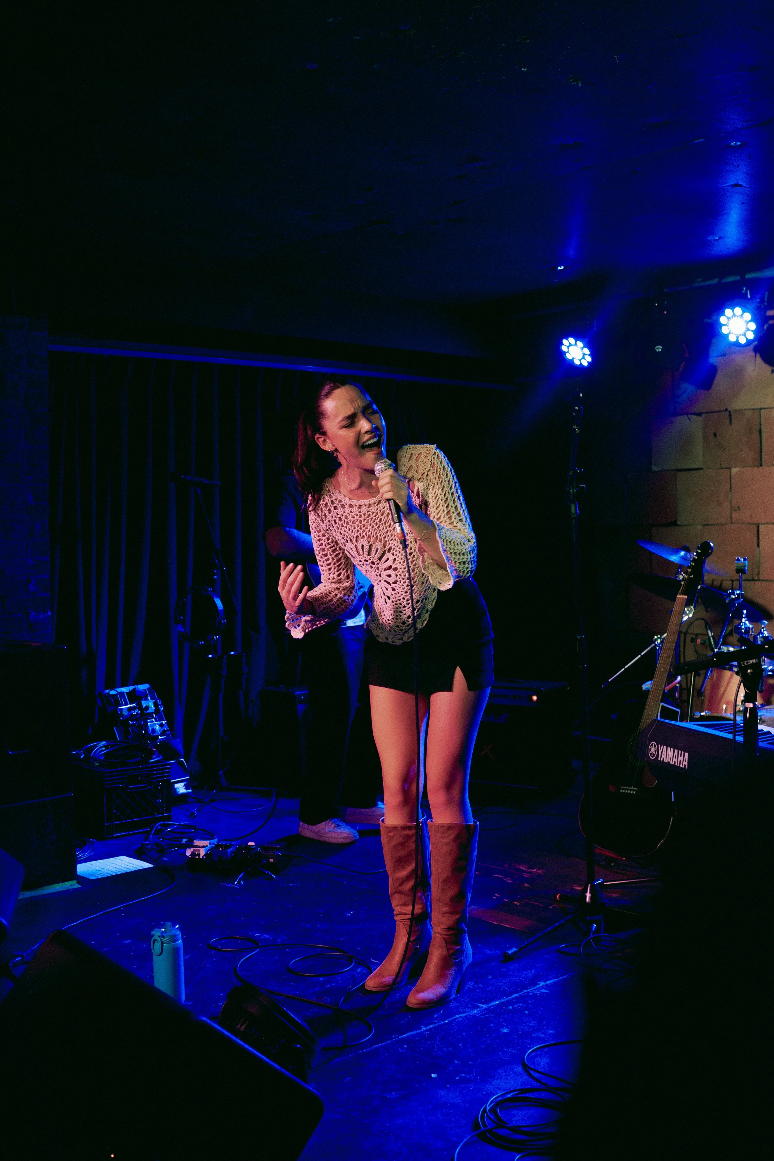A woman singing passionately into a microphone on stage during a live music performance, with blue stage lights illuminating the scene.