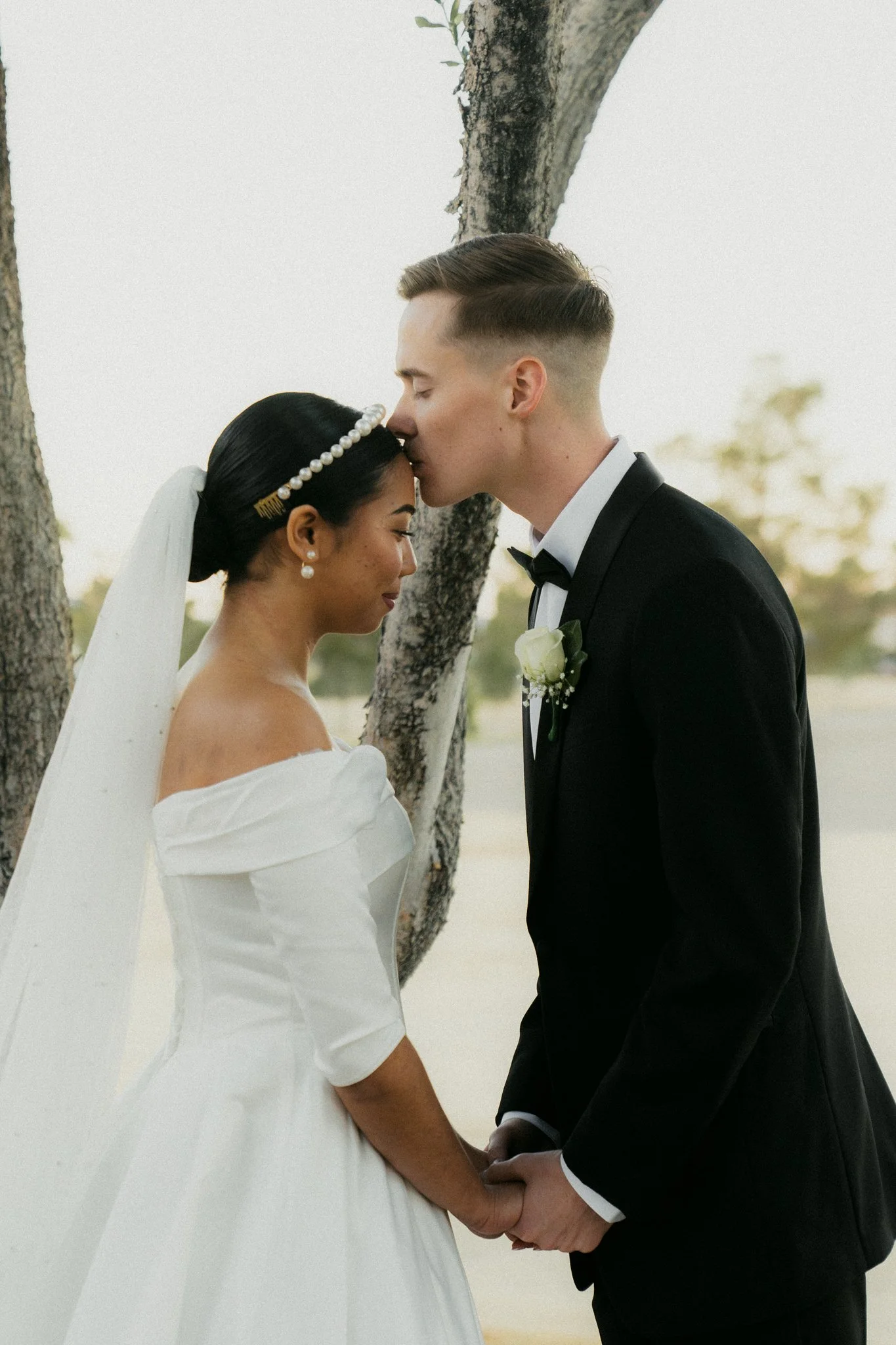 A bride and groom sharing a romantic moment outdoors, holding hands in front of a tree. The bride is wearing a white off-shoulder wedding dress, pearl hair accessories, and a veil. The groom is wearing a black tuxedo with a white shirt and black bow 