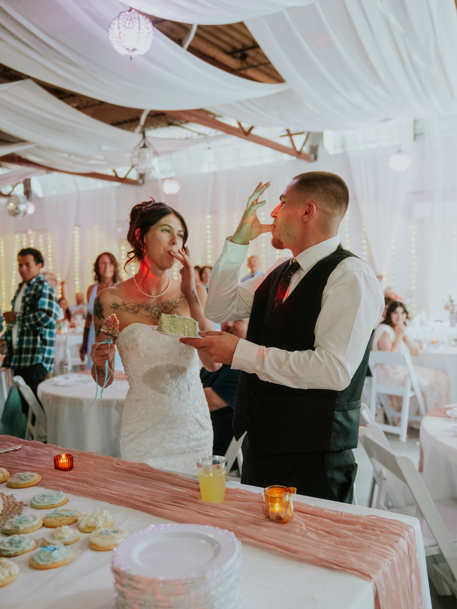 A bride and groom at their wedding reception, with the bride wearing a white lace wedding dress and the groom in a black vest and white shirt, sharing a moment with the bride eating cake.