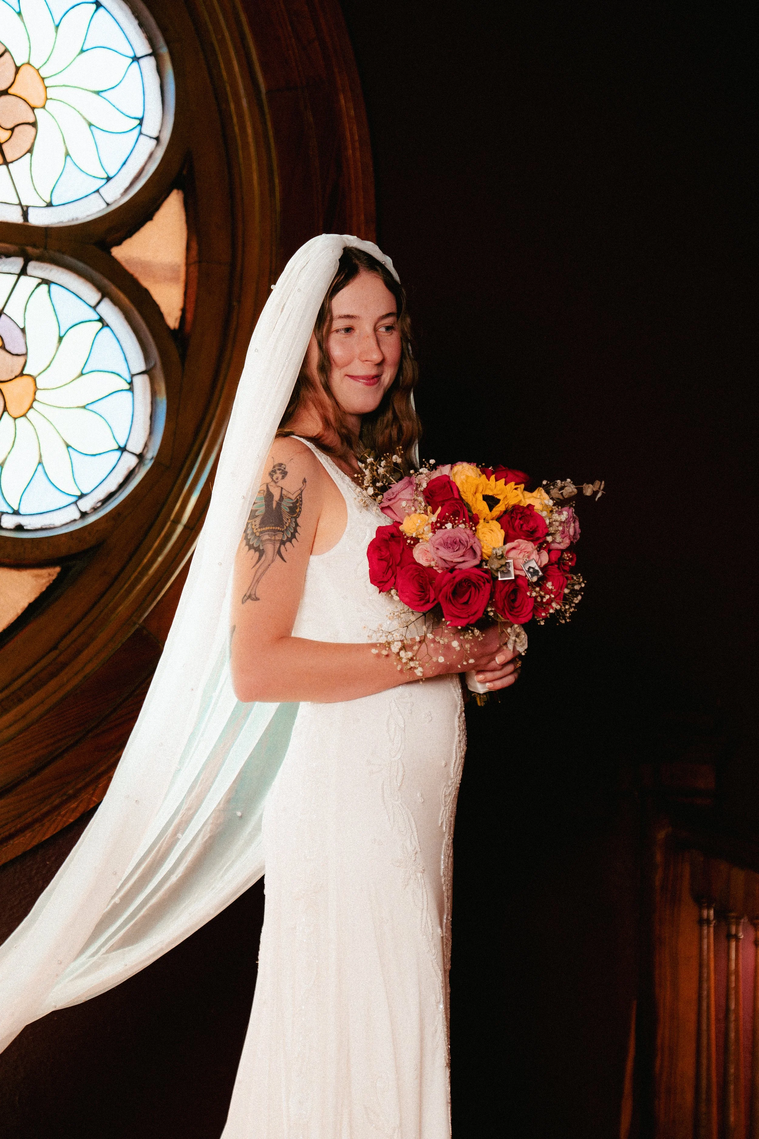 A bride in a white wedding dress holding a bouquet of red, pink, and yellow flowers, standing inside a church with stained glass windows.