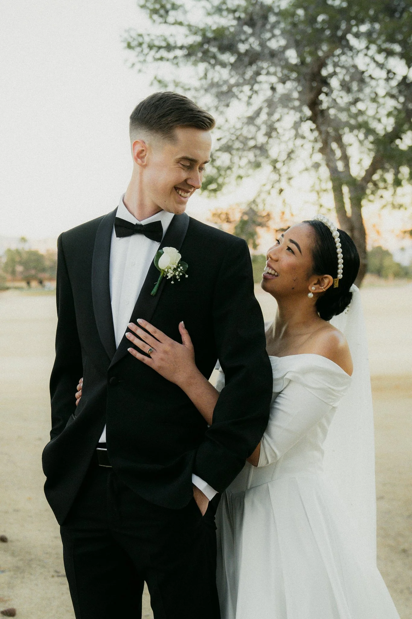 A smiling bride and groom share a moment outdoors, the bride in a white wedding dress and the groom in a black tuxedo, standing close with the bride touching the groom's chest.