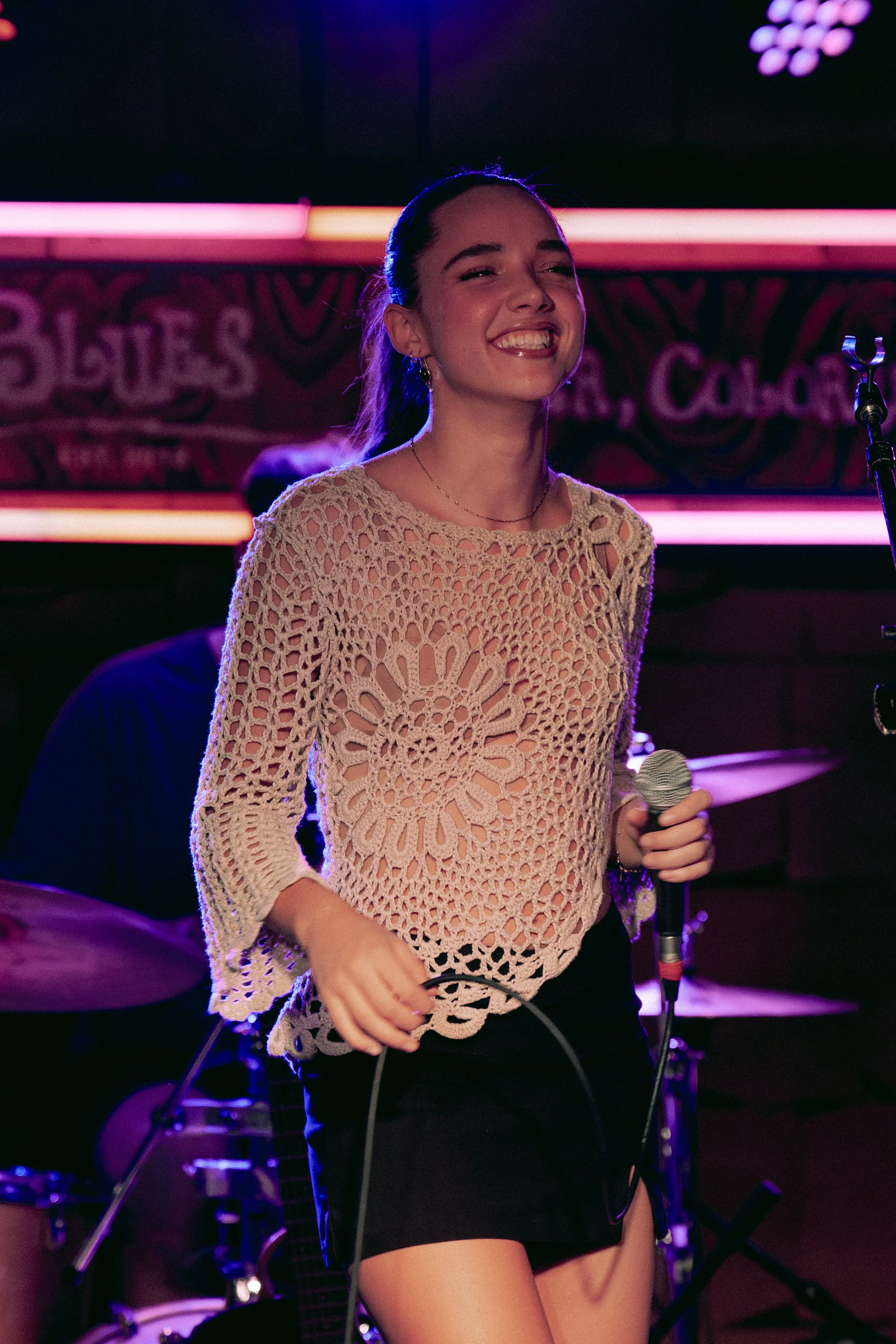 A young woman with long hair tied back, smiling and holding a microphone, standing in front of a colorful stage background with neon lights, dressed in a crochet top and black shorts.
