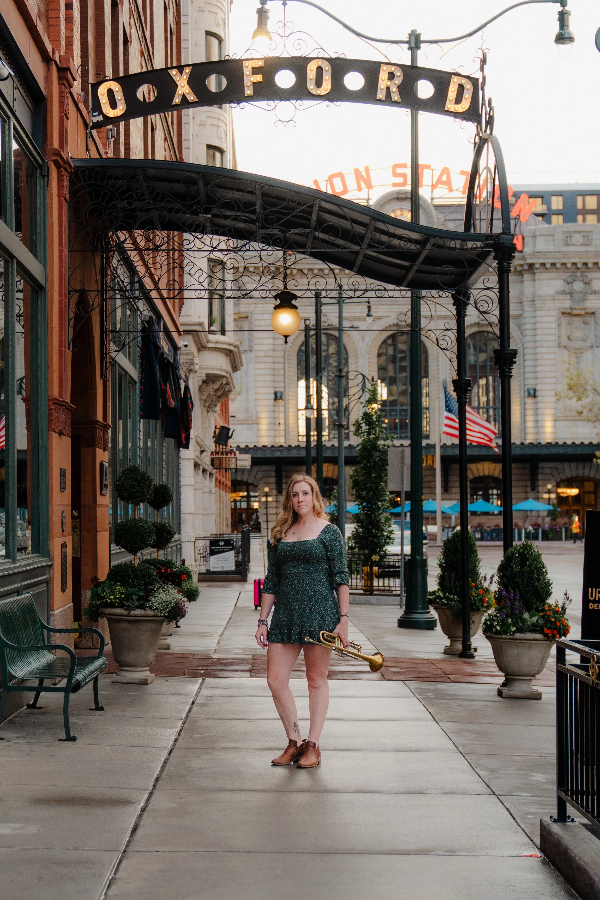 A young woman standing on a city sidewalk at dusk, holding a trumpet, surrounded by planters with flowers, with historic buildings and neon signs in the background.