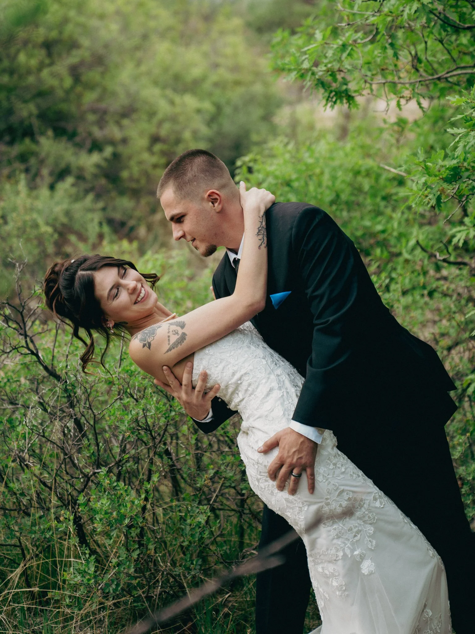 A newlywed couple dancing outdoors among greenery, with the groom in a black tuxedo and the bride in a white lace dress, sharing a joyful moment.