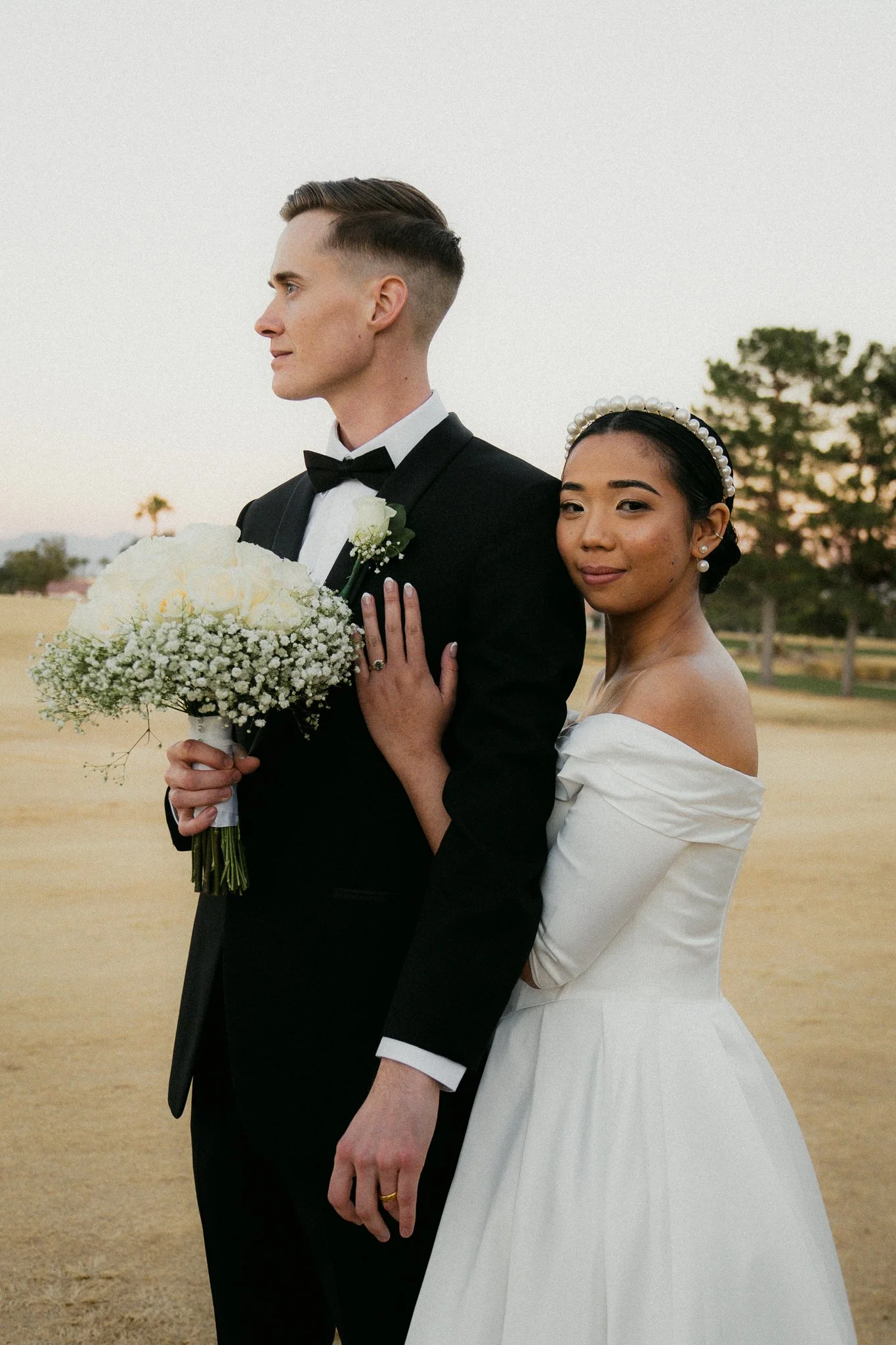 A couple in wedding attire standing outdoors; the groom in a black tuxedo holding a bouquet of white flowers, and the bride in a white off-shoulder gown wearing a pearl headband.