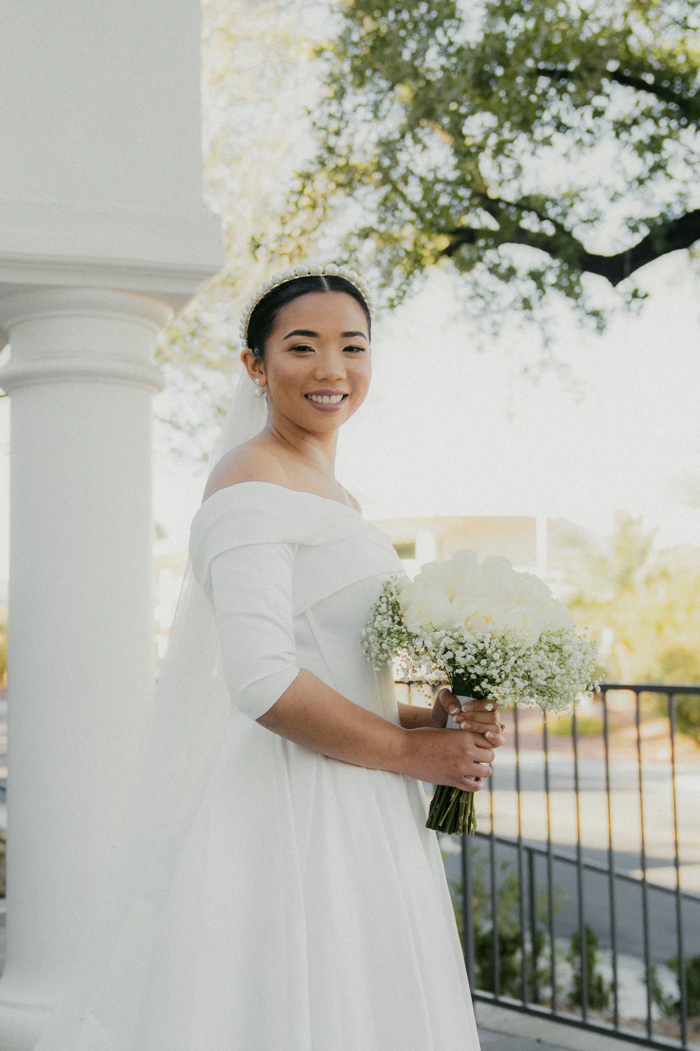 A smiling bride in a white wedding dress holding a bouquet of white roses and baby's breath, standing outdoors near a white column and a railing, with trees in the background.